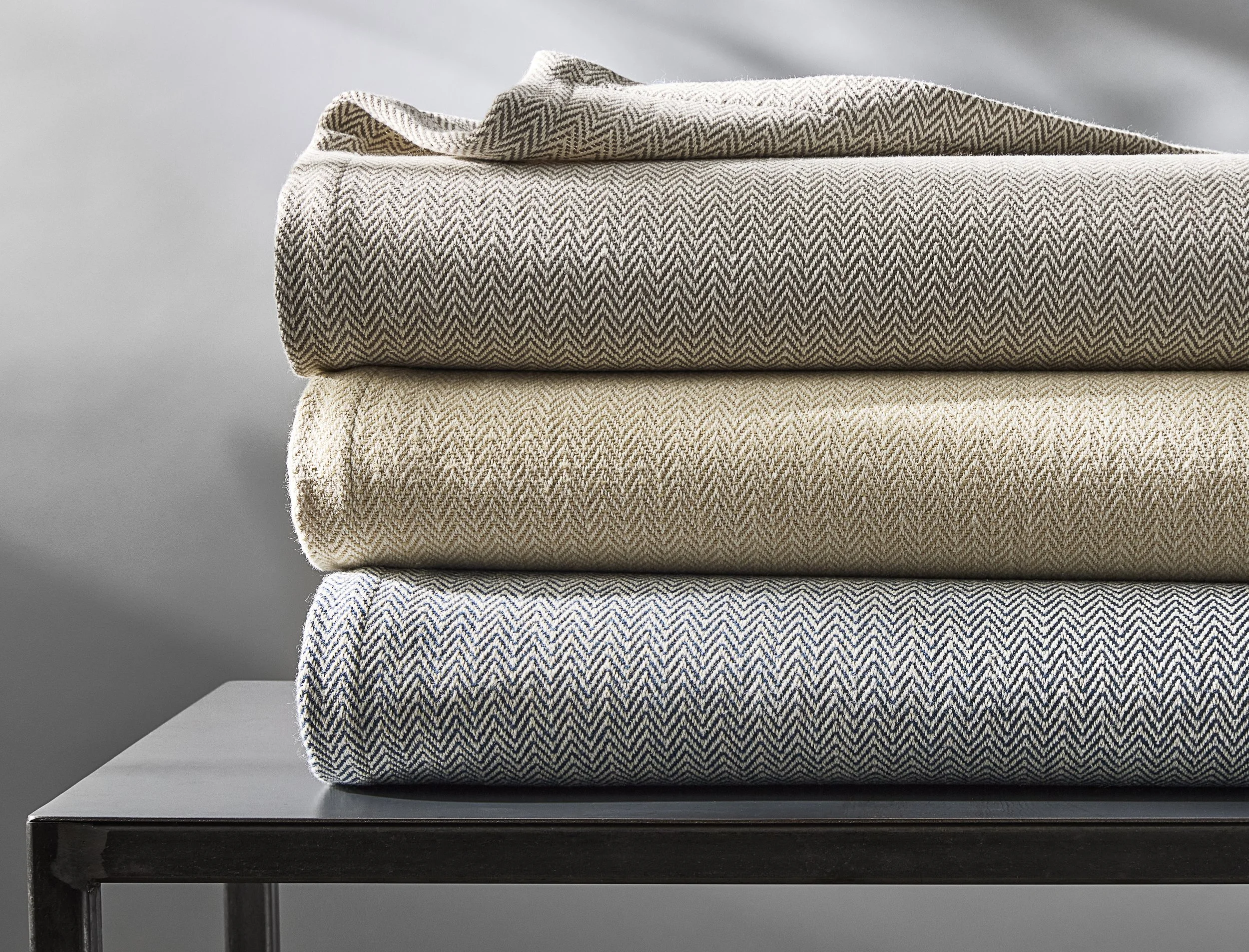 Three folded textiles with zigzag patterns in beige, cream, and navy are stacked on a black table against a gray background.