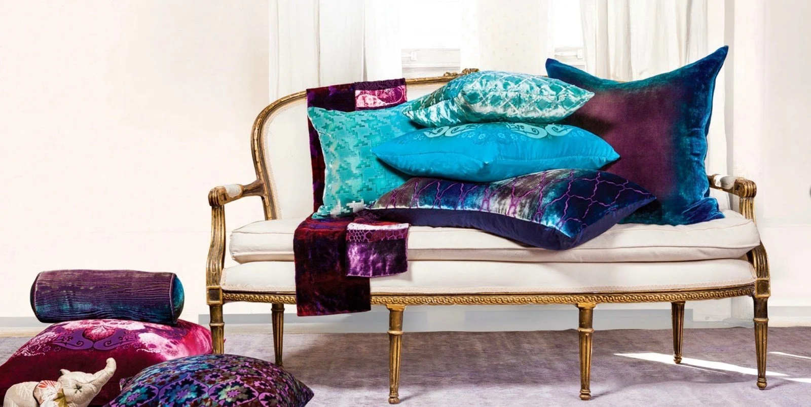 A vintage white sofa with a gold frame is decorated with multiple colorful velvet pillows in shades of blue, teal, purple, and patterned fabrics, with additional pillows and fabric pieces on the floor nearby, set against white curtains and walls.