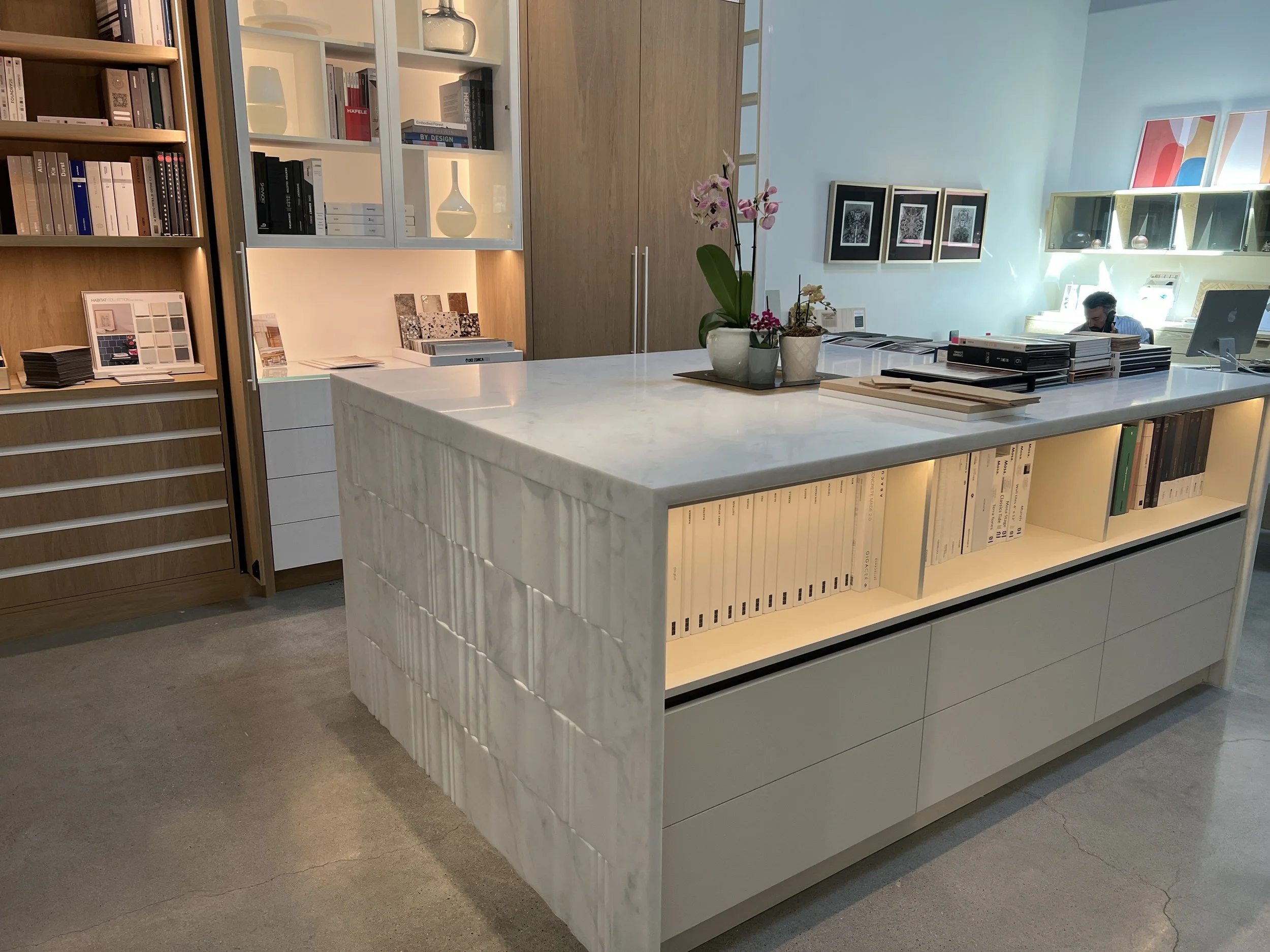 Modern office space with a large white marble island counter, illuminated shelf storage underneath, and various books and decorative items on shelves and in the background. A person is sitting at a desk in the distance.