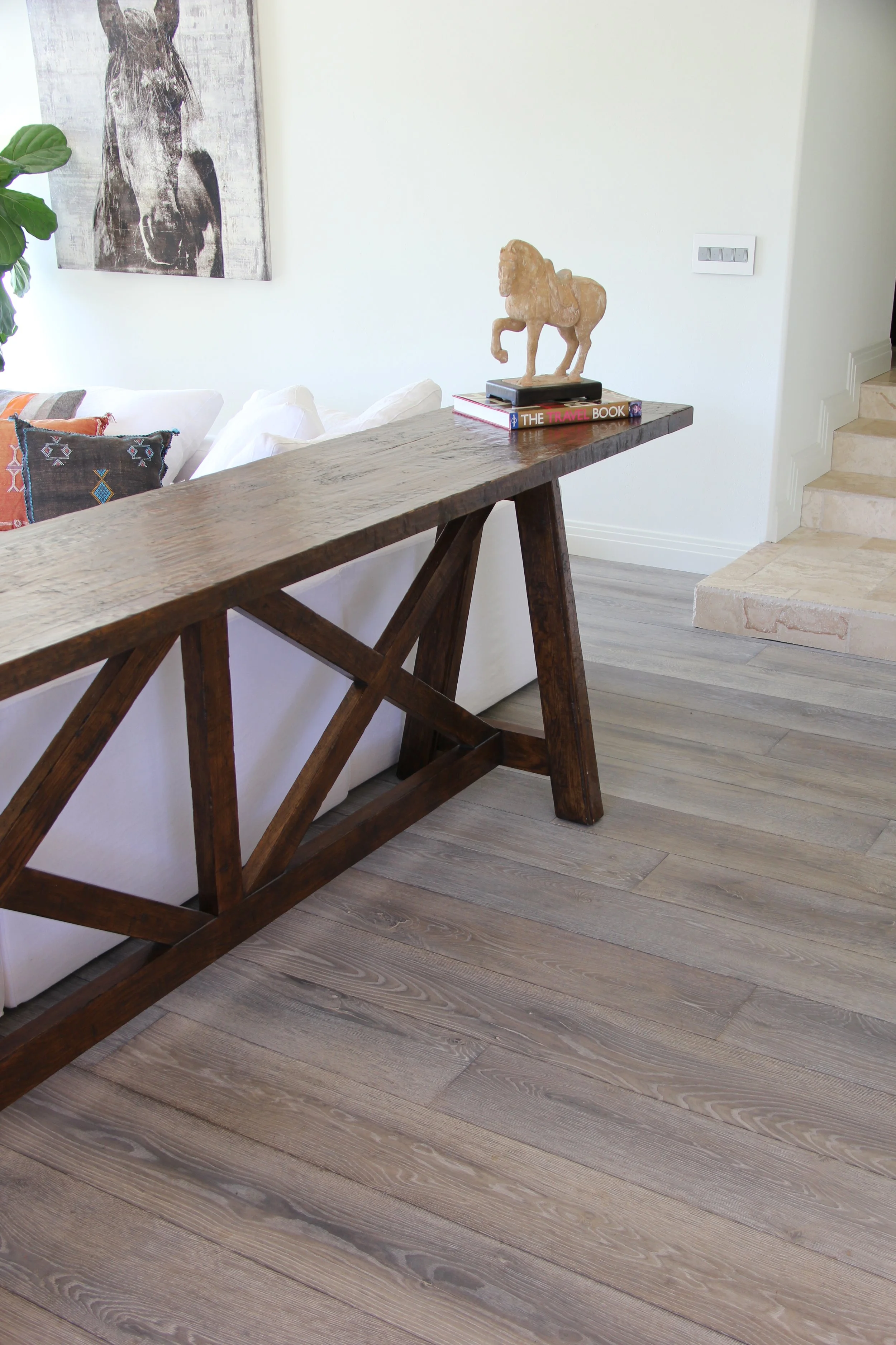 Living room with a wooden console table, a white sofa with colorful pillows, a tribal horse sculpture on top of books, and a black and white horse painting on the wall.