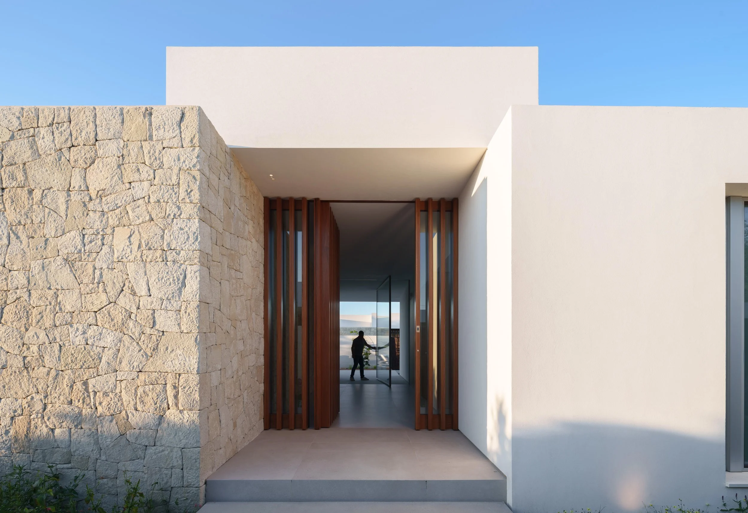 Modern house with stone and white exterior walls, a wooden entrance door, and a person inside visible through glass panels.