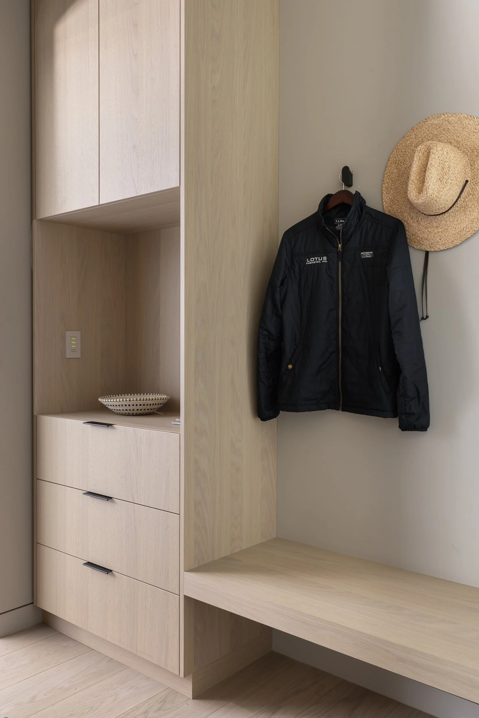 A wooden entryway with a black jacket hanging on a hook, a straw hat, a wooden bench, and drawers.