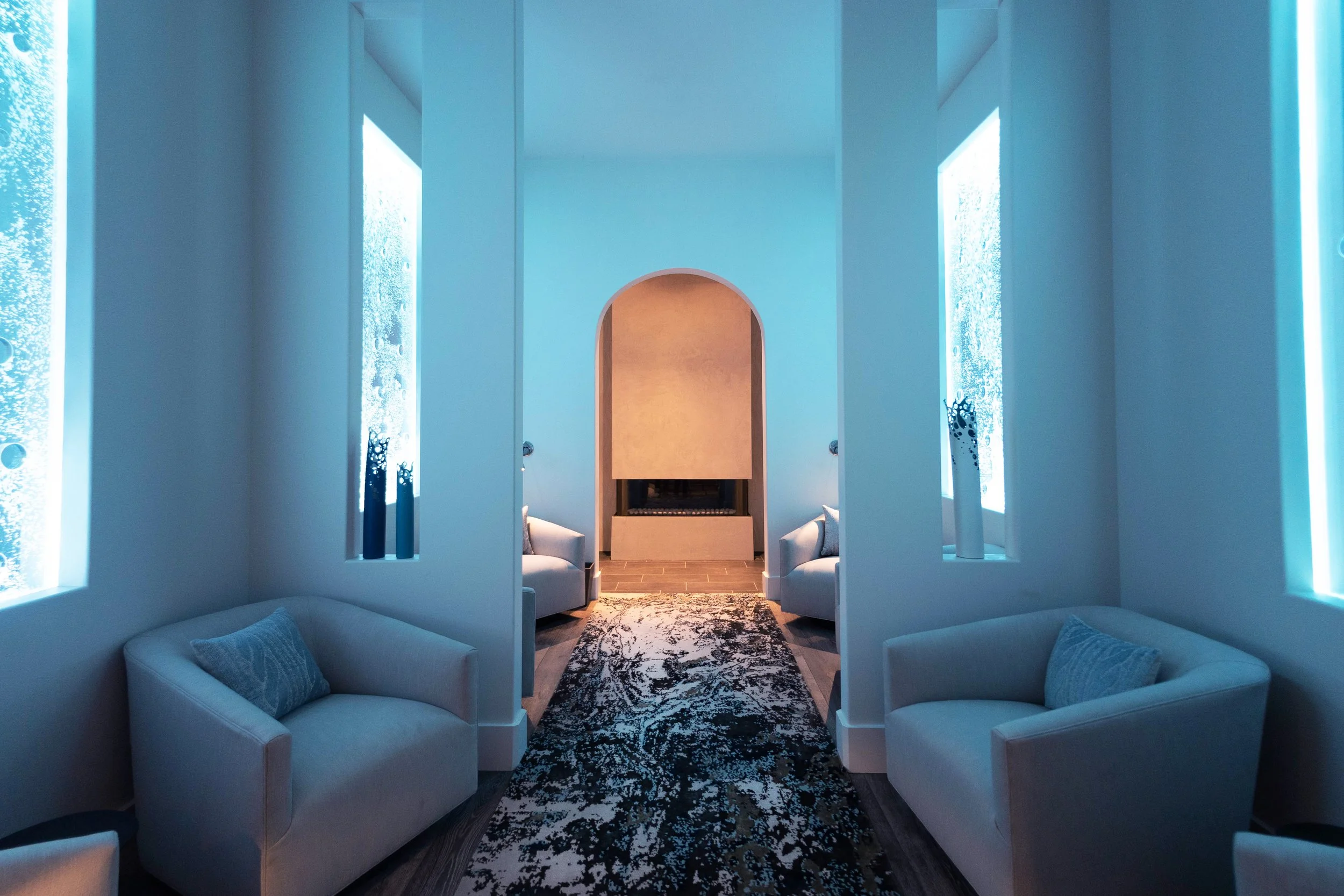 A modern living room with white armchairs, a black and white patterned rug, large windows with blue-tinted glass, and a fireplace at the back, all painted in soft white and blue tones.