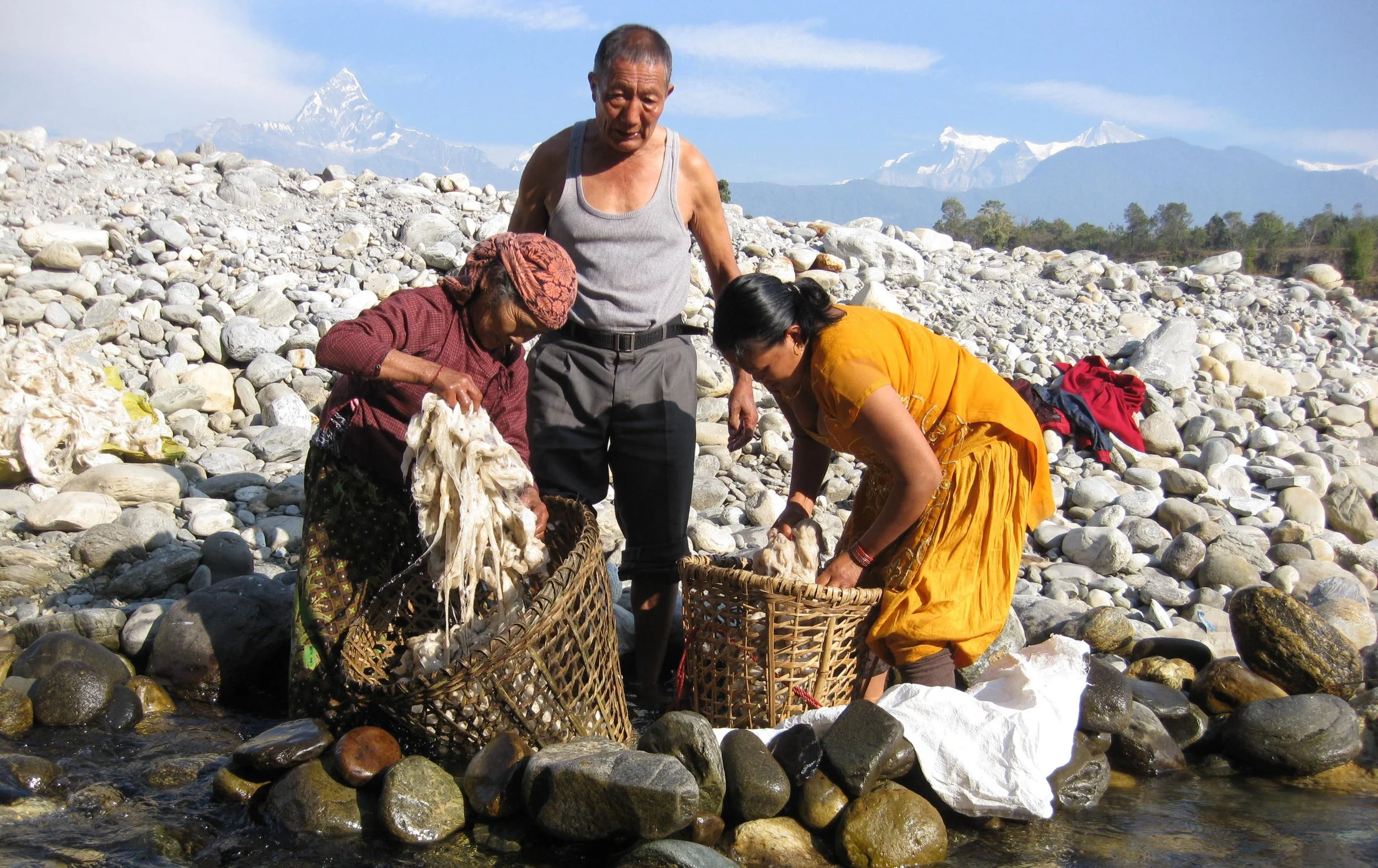 Three people, two women and one man, collecting wool or fiber from a rocky stream in the Himalayas with snow-capped peaks in the background.
