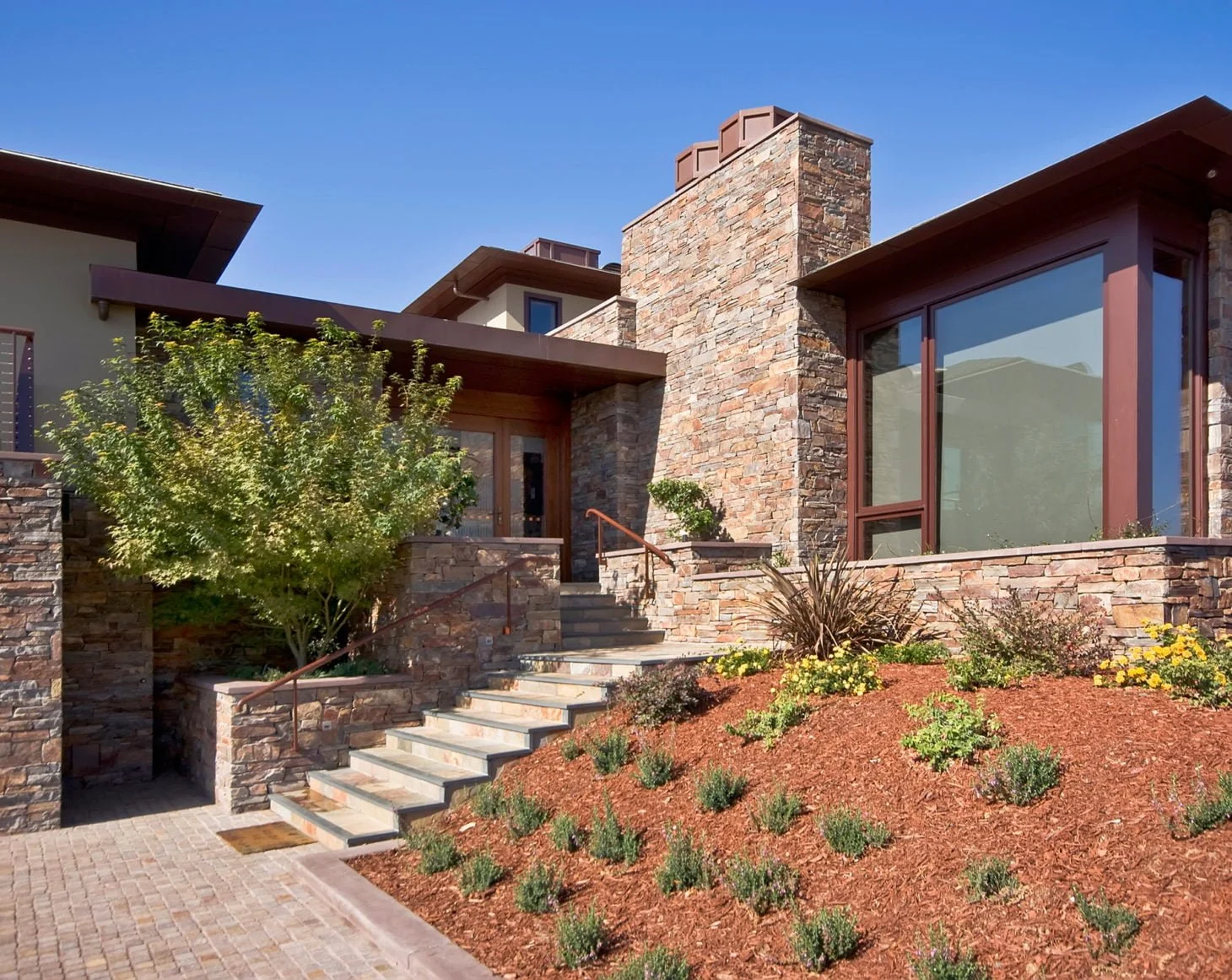 Exterior view of a modern home with stone and glass facade, landscaped garden, and cobble stone driveway.