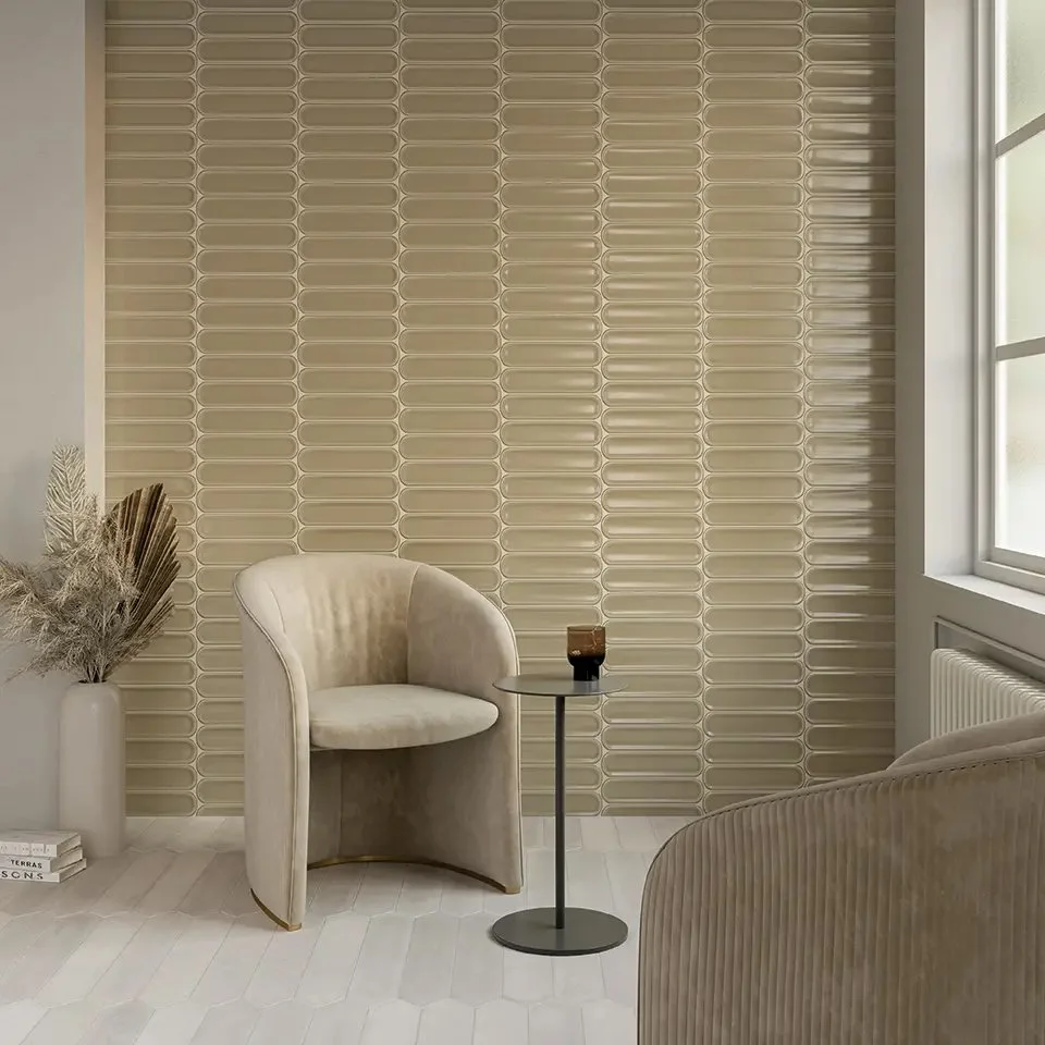 A cozy corner with two beige upholstered chairs, a small black side table holding a brown cup, a tall white vase with dried neutral-tone ornamental grasses, a stack of books, and a textured tile wall in a neutral beige color beside a window.