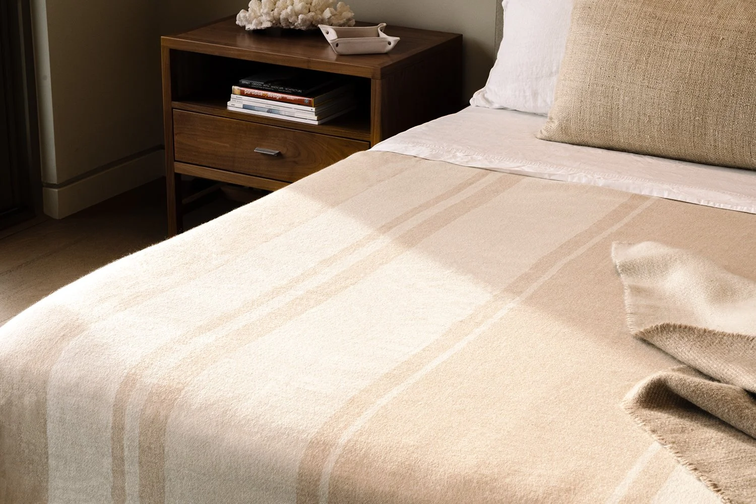 A neatly made bed with beige bedding and pillows, beside a wooden nightstand with books and decorative items, in a cozy bedroom.