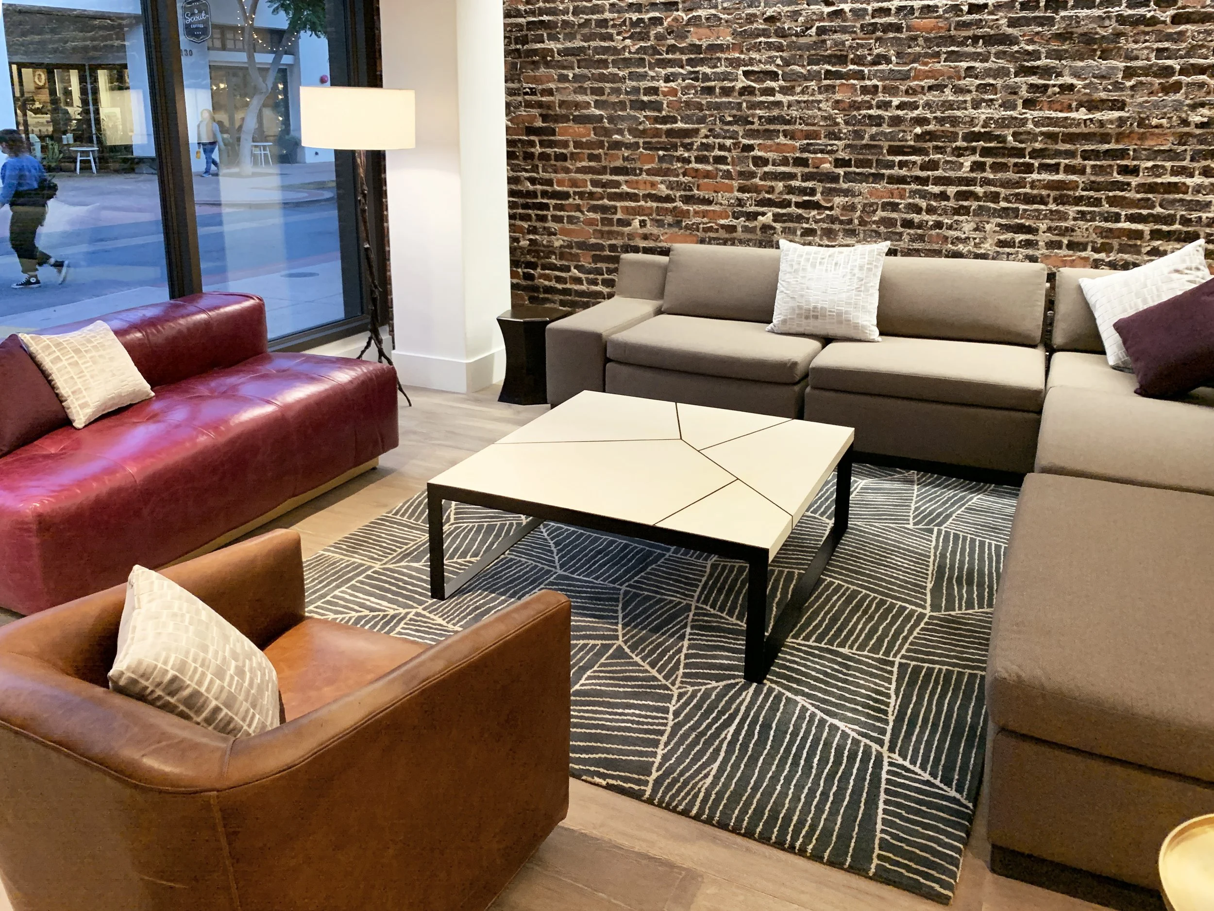 Lounge area with a red leather sofa, a tan sectional sofa with decorative pillows, a brown leather armchair with a pillow, a geometric patterned rug, a modern white coffee table, a tall floor lamp, and a large window revealing pedestrians outside.