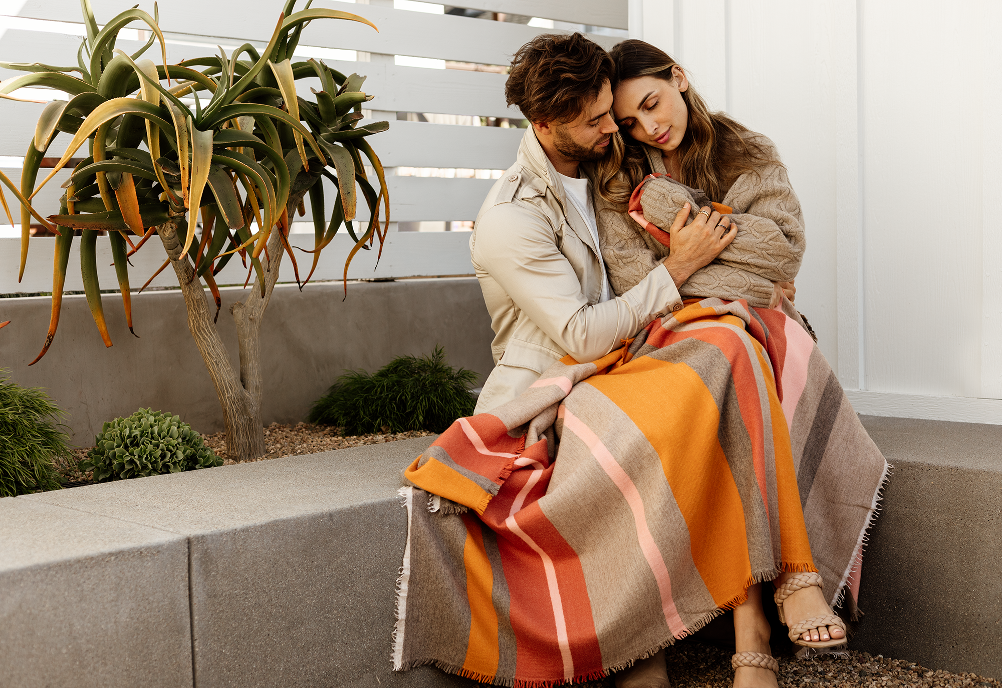 A couple sitting outdoors on a concrete bench, wrapped in an orange, pink, and gray plaid blanket, holding a small puppy, with plants and a white wooden fence in the background.