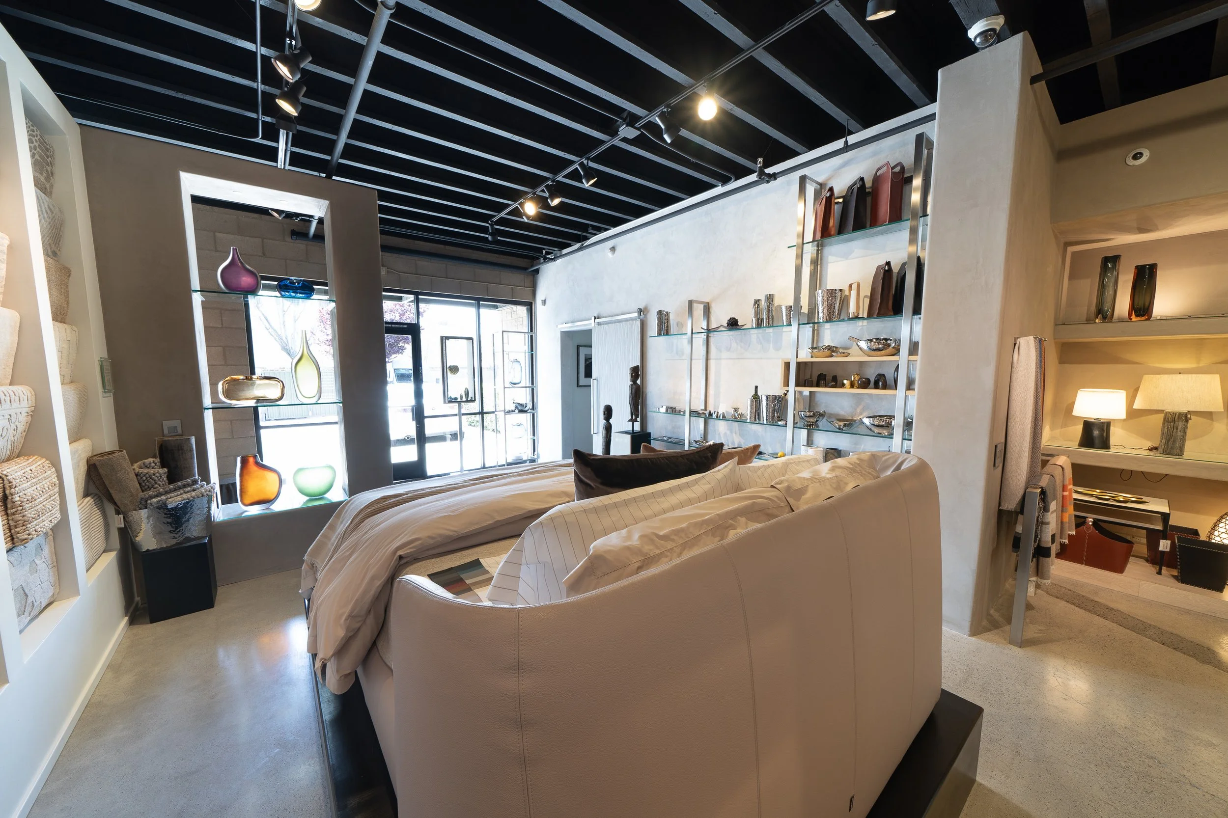 Interior of a modern furniture and home decor store with a white couch, shelves with vases and decorative items, large windows, and ceiling track lighting.