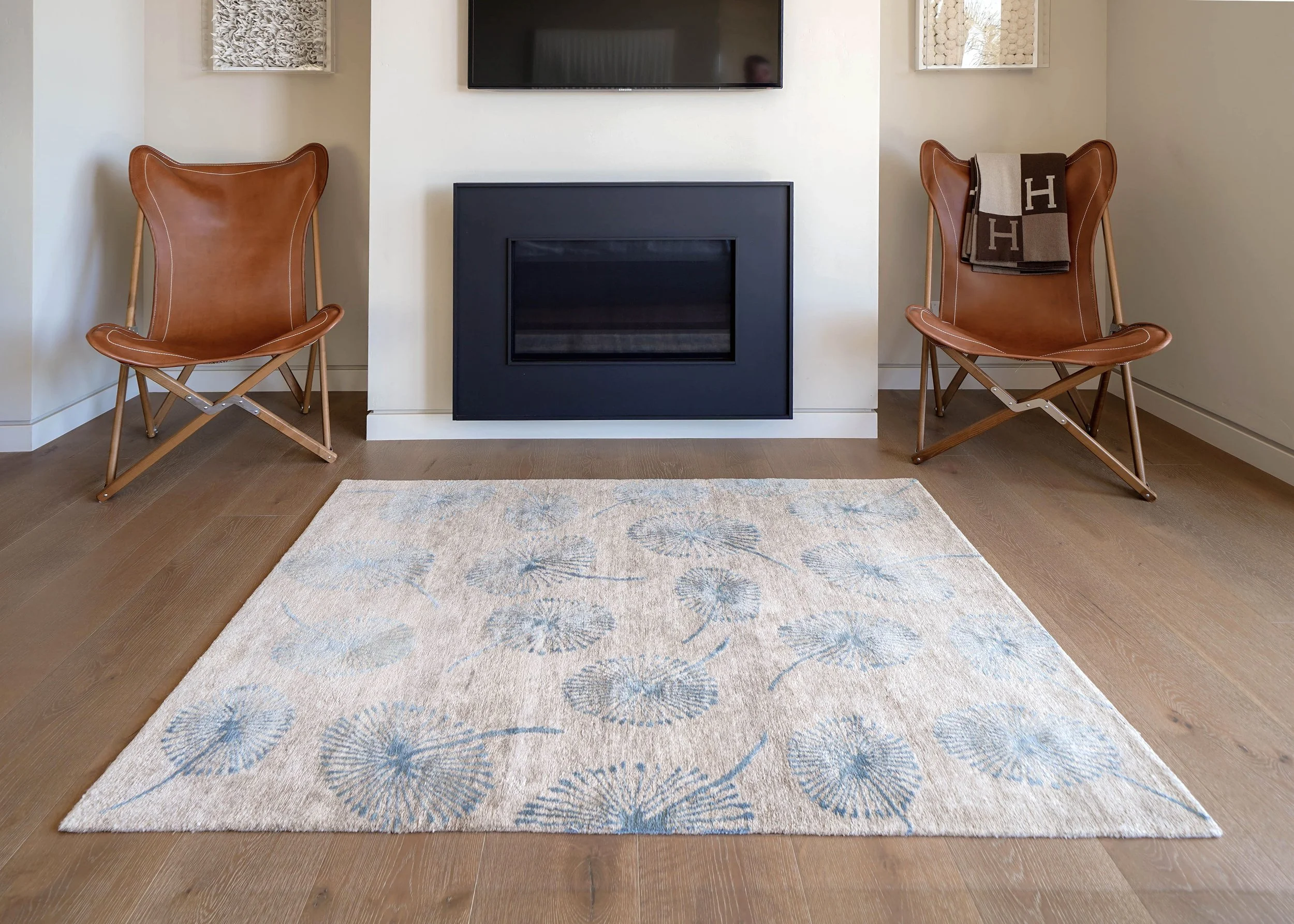 Living room with two brown leather and wood framed chairs, a white wall with a mounted black TV above a black fireplace. A beige rug with blue floral patterns is on a wooden floor.