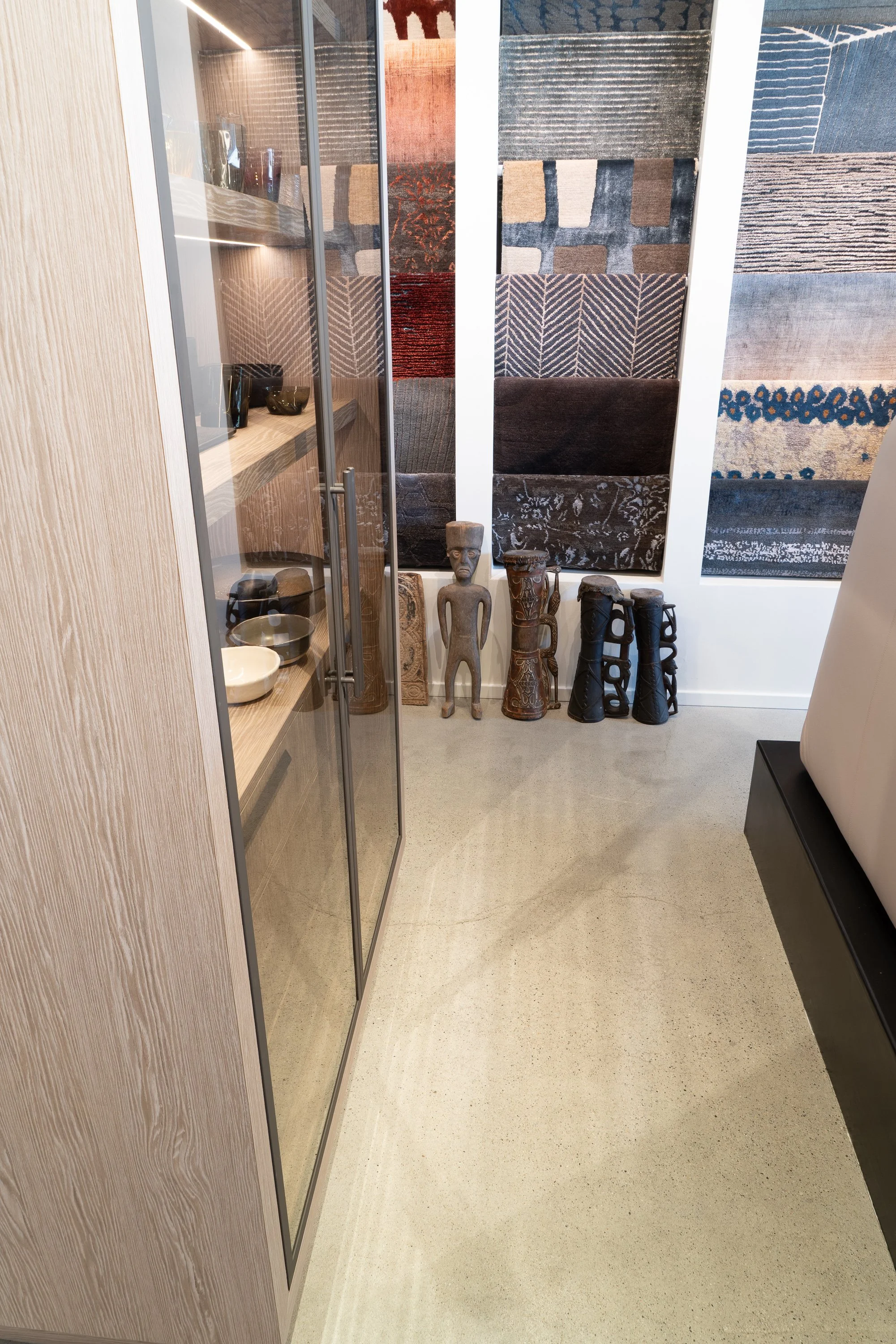 Display of various rugs on the wall, sculptures and artifacts on the floor, a wooden cabinet with glass doors on the left, and a portion of a bed on the right, in a room with a polished concrete floor.