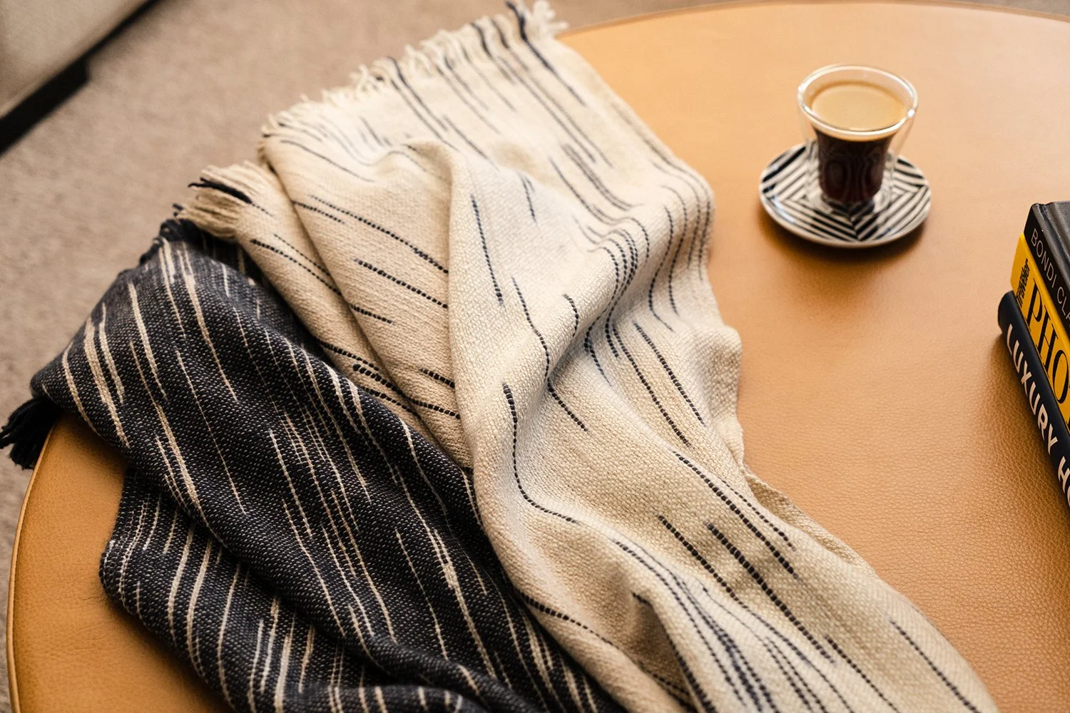 Two striped scarves, one black and white and the other beige with black lines, lying on a tan table next to a cup of coffee on a saucer, a stack of books, and a beige carpet.