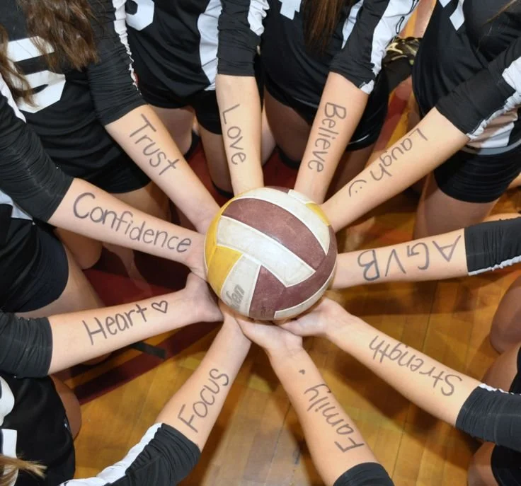 A group of volleyball players in black uniforms with white stripes on their sleeves, standing in a circle with their arms extended inward, each hand touching a volleyball at the center. Words are written on their arms including 'Trust,' 'Love,' 'Believe,' 'Laugh,' 'Courage,' 'Strength,' 'Focus,' 'Heart,' 'Confidence,' and 'Achieve.'