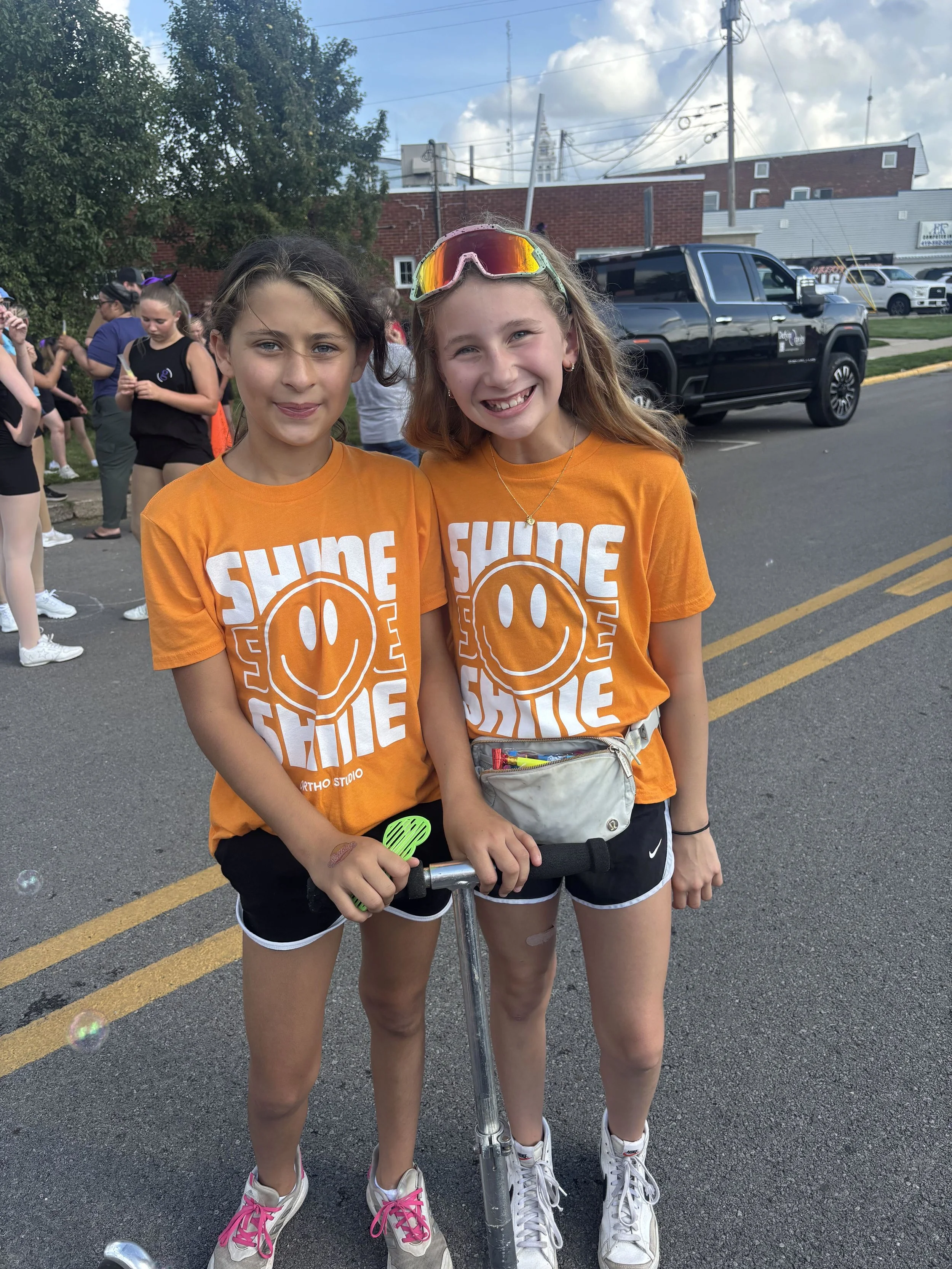 Two smiling young girls wearing orange Shine Ortho Studio shirts standing with scooters during community parade