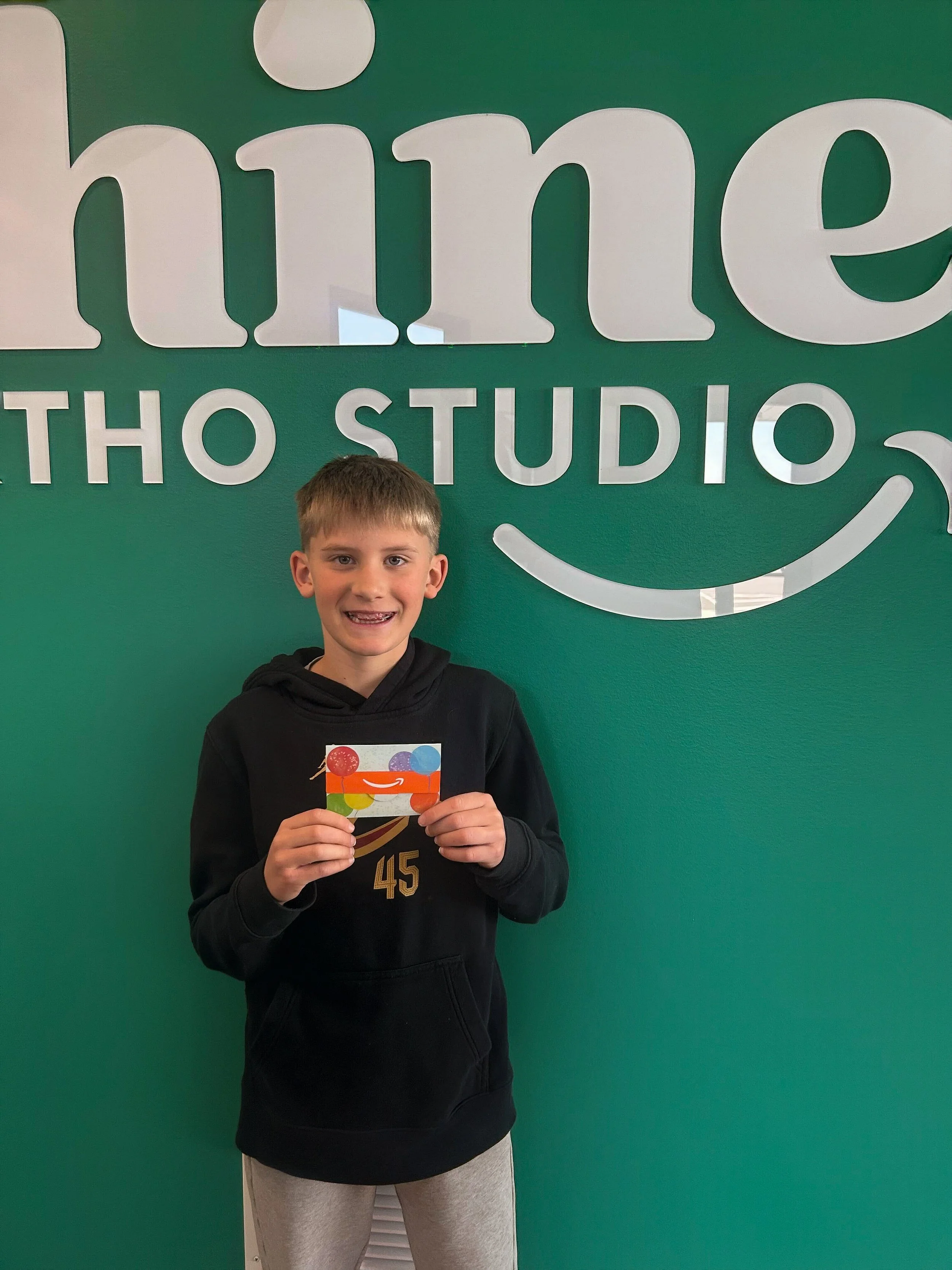 Child smiling and holding a gift card in front of the Shine Ortho Studio sign, representing a referral or rewards program winner.
