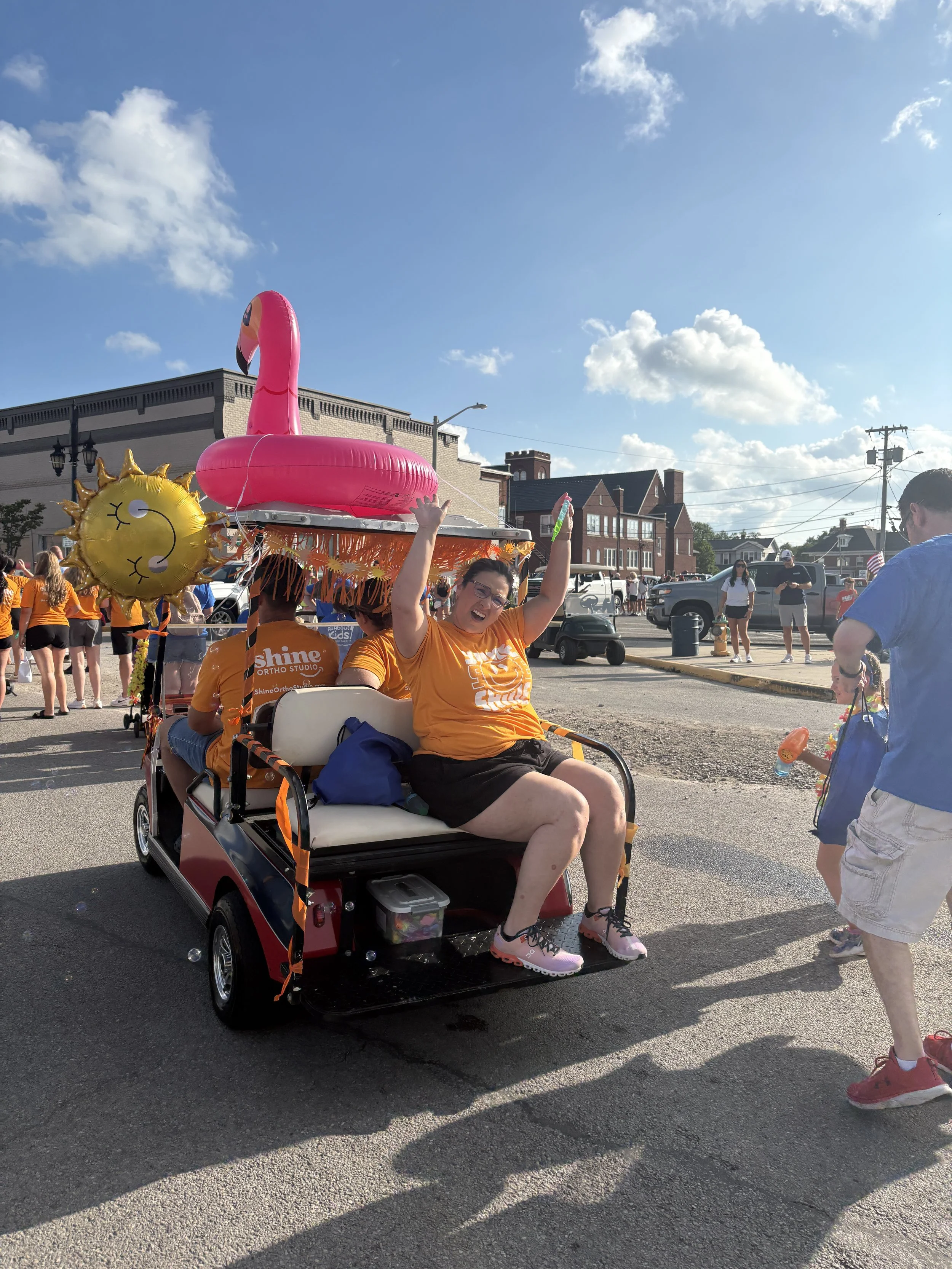 Shine Ortho Studio team member celebrating on decorated golf cart float with pink flamingo and smiley sun balloon at parade