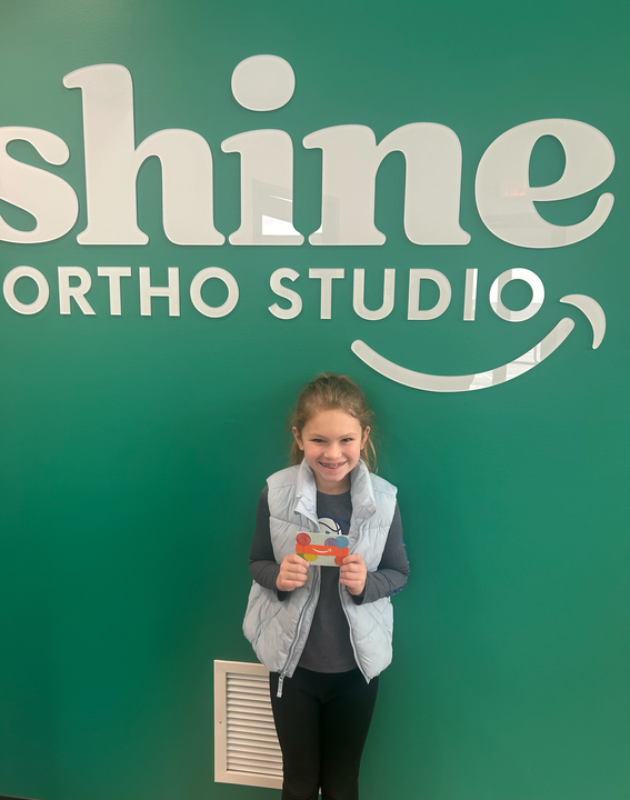 Young patient smiling and holding a gift card in front of the Shine Ortho Studio wall logo, representing a referral reward winner.