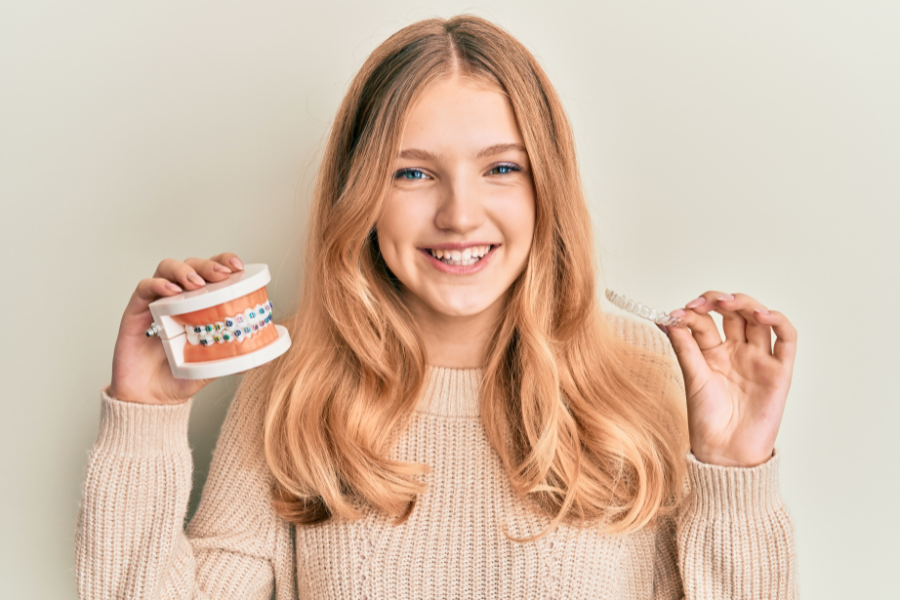 Teen girl smiling while holding a braces model and a clear aligner tray, representing orthodontic treatment options at Shine Ortho Studio.