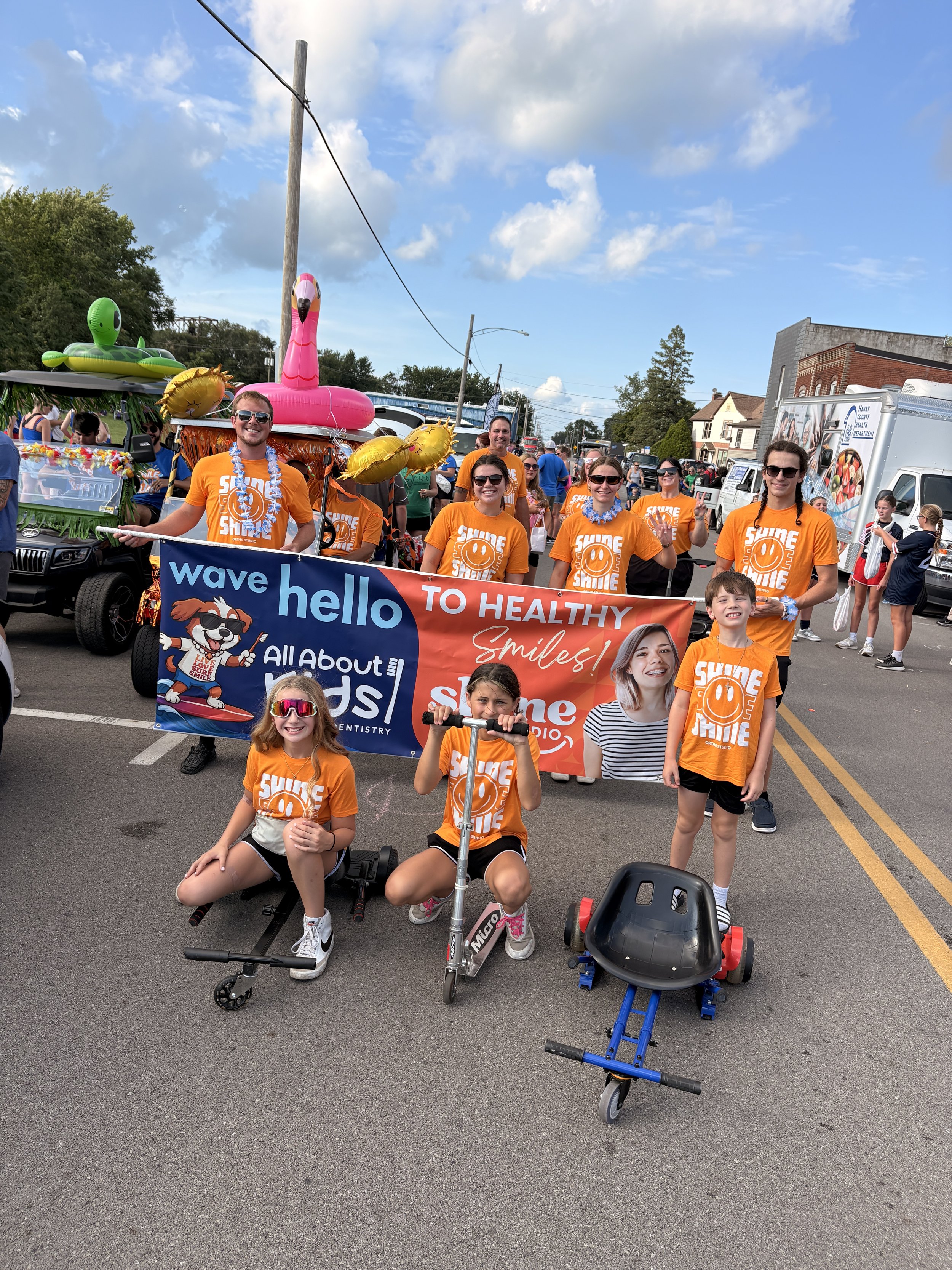 Shine Ortho Studio team and children on scooters posing with parade banner that reads wave hello to healthy smiles in front of decorated float