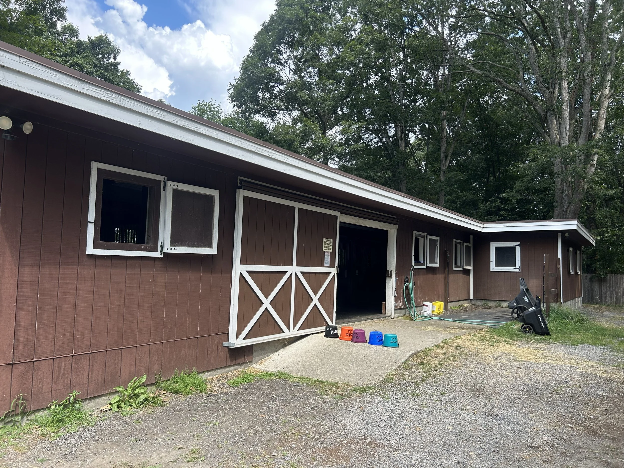 Brown barn with open windows and a sliding door, surrounded by trees, with buckets and cleaning equipment outside.