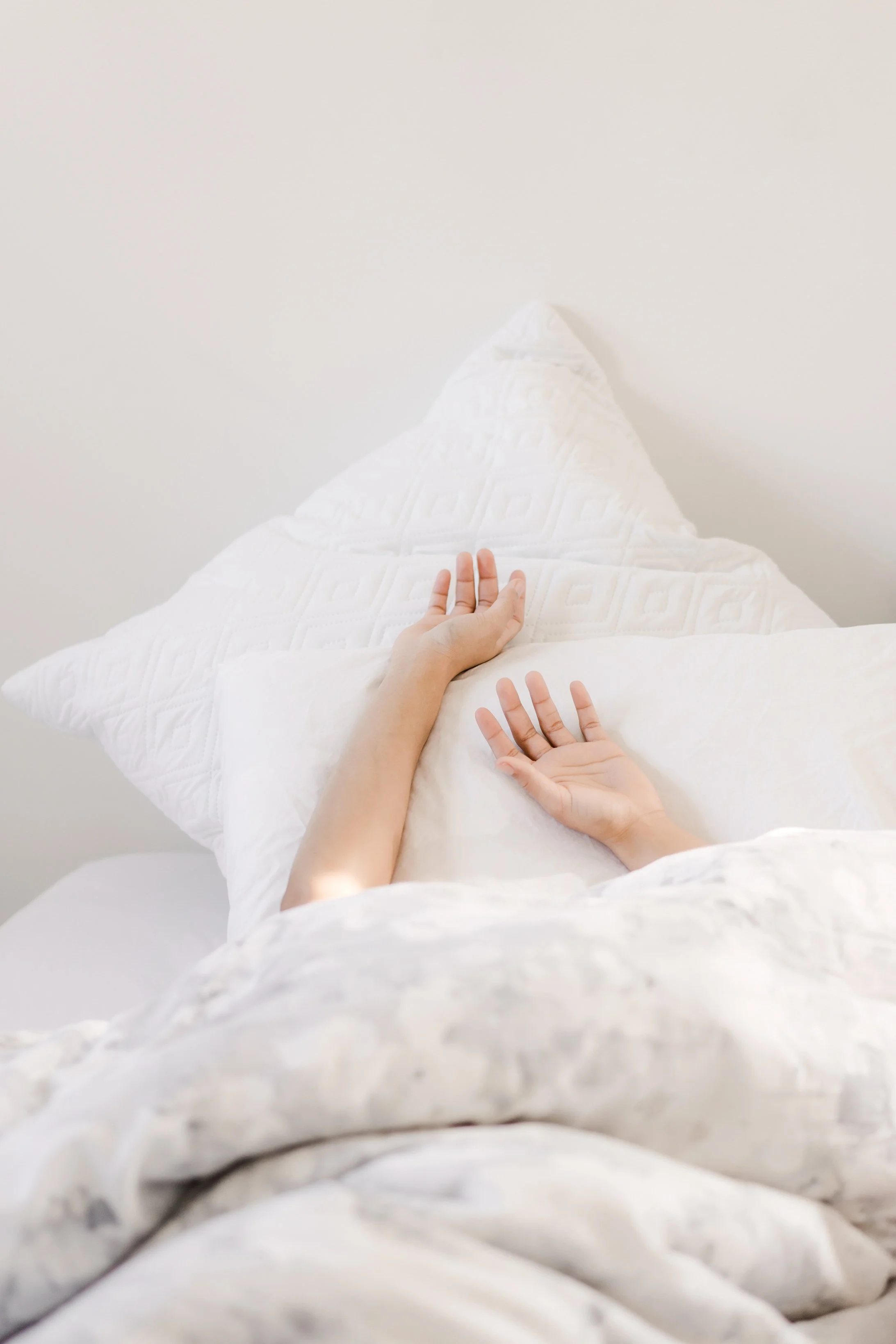 Person's hand resting on a white pillow on a bed, with white blankets and bedding, against a plain white wall.