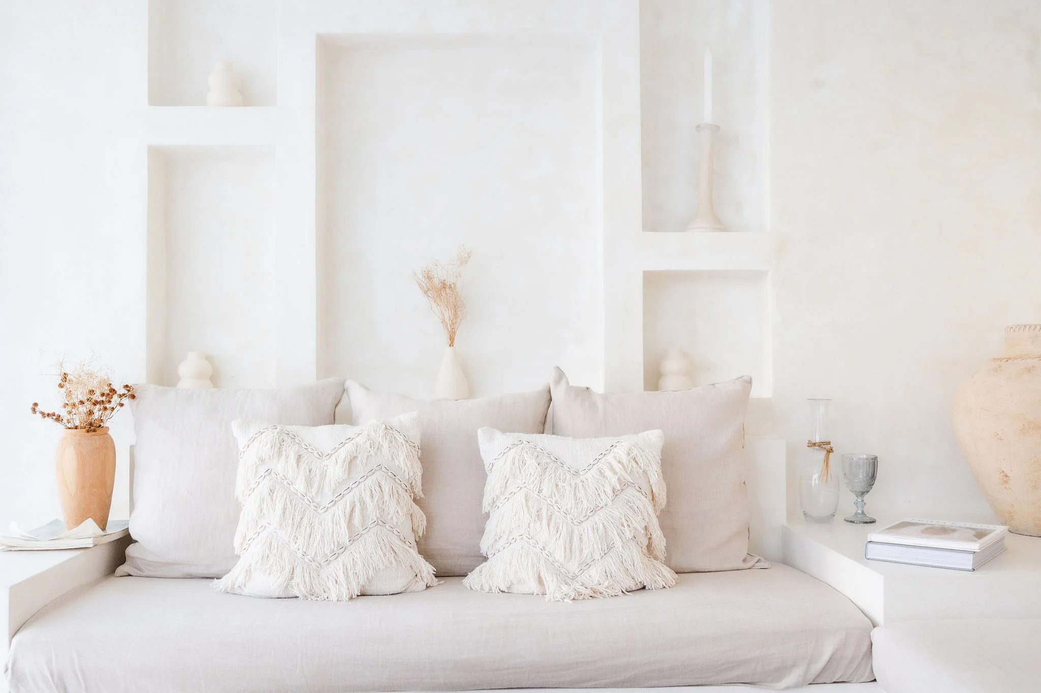 A white sofa with multiple textured white pillows in front of a white wall with built-in niche shelves decorated with vases and a candle, flanked by plants in vases and glasses on a side table.