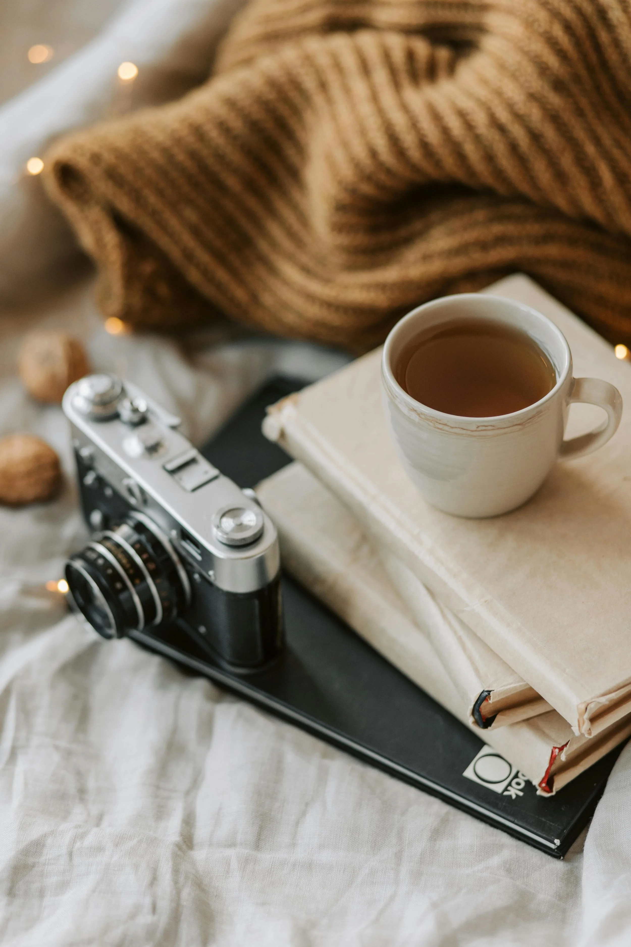 A cozy scene with a cup of tea on top of a stack of books, a vintage camera, a brown knit blanket, walnuts, and a black notebook on a white fabric surface. Self-care, creative, journalling invitation.