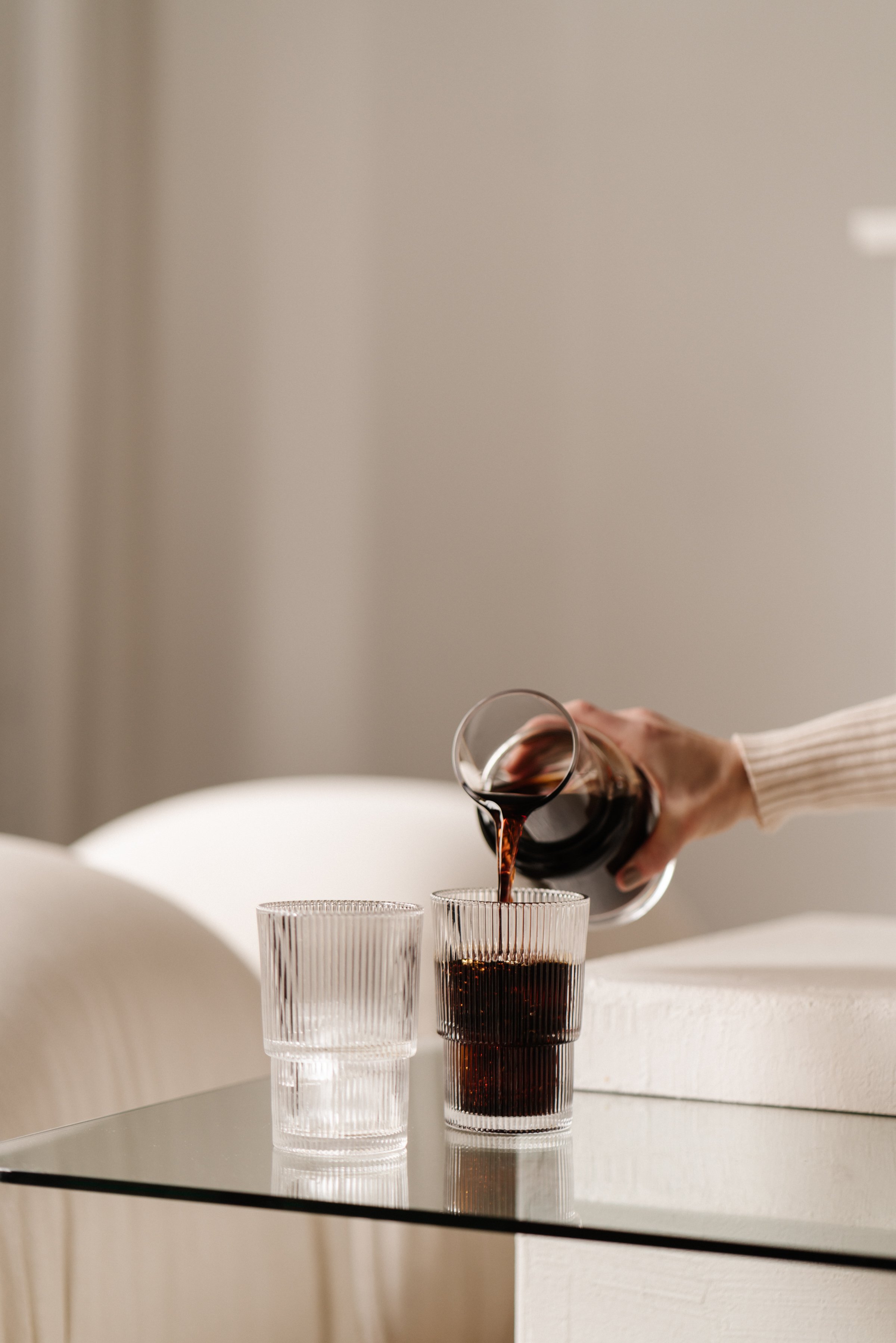 Person pouring red wine into a glass on a table with two other empty glasses, with a neutral beige wall in the background.