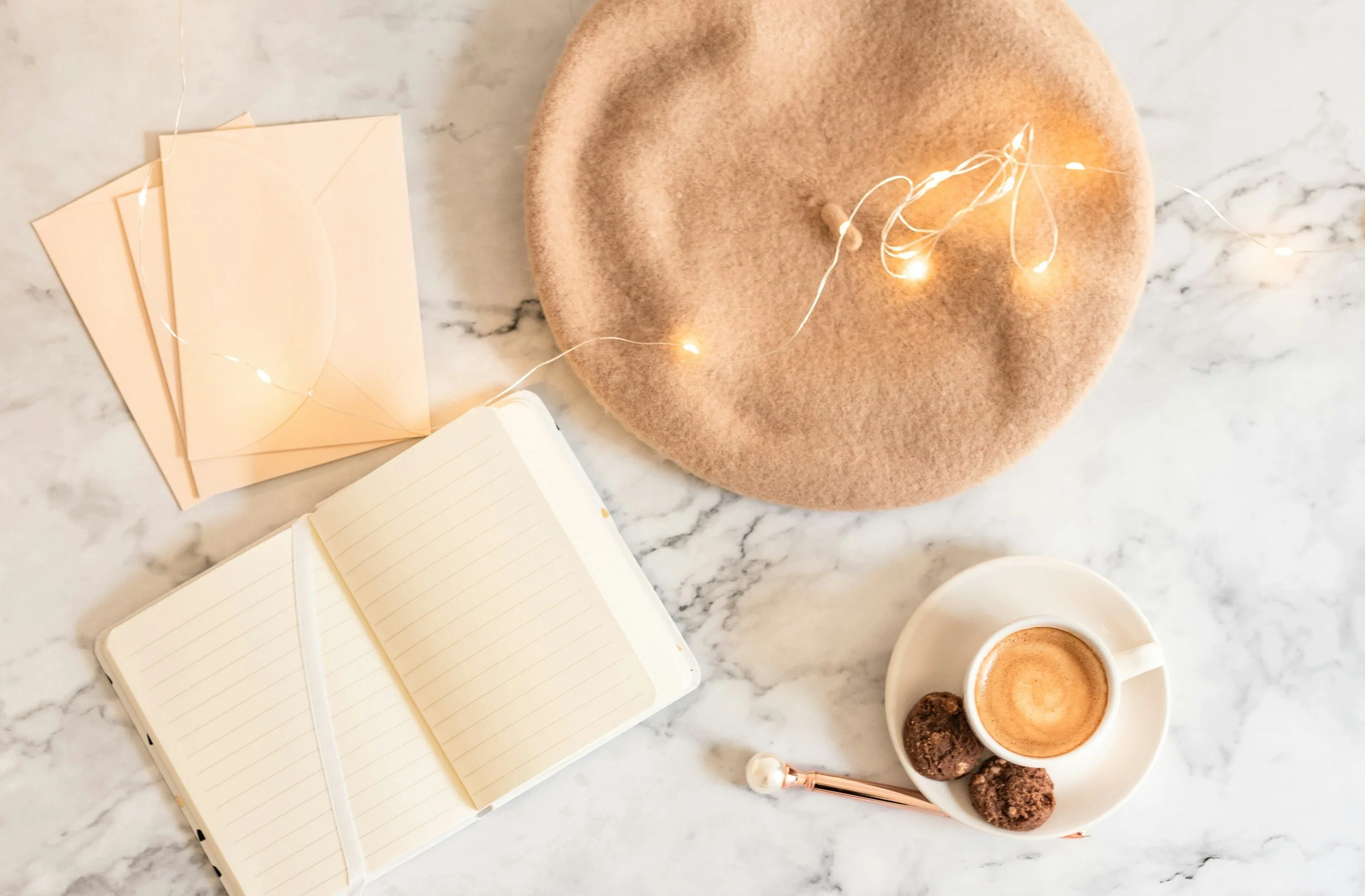 Items on a marble table, a beret, a string of lights and notebook and a coffee with two brown cookies.