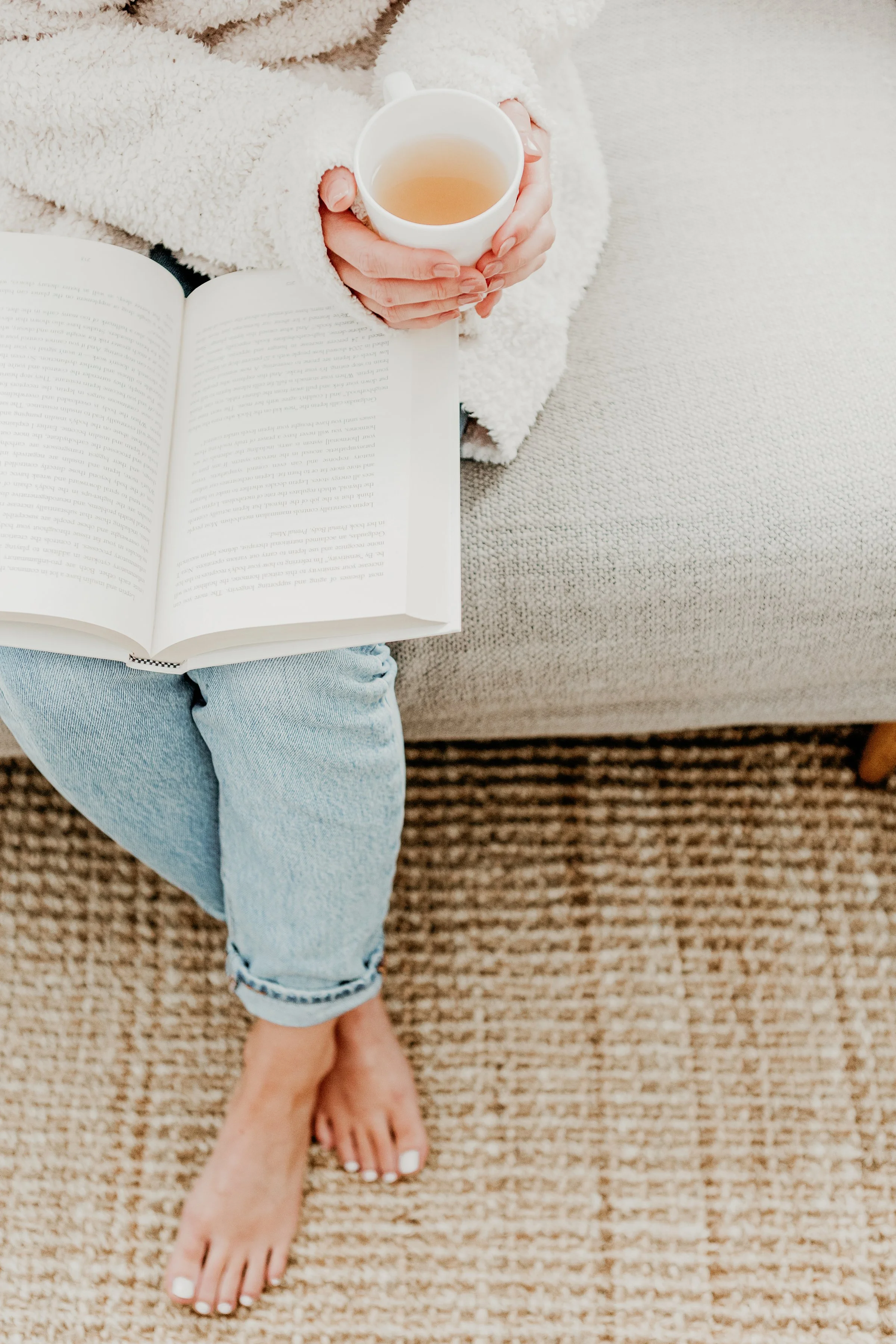 Person sitting on a beige carpet, holding a white mug of tea with both hands, wearing a soft, fluffy white sweater. There is an open book on their lap, and their legs are crossed at the ankles, wearing light blue jeans. Their toenails are painted white.