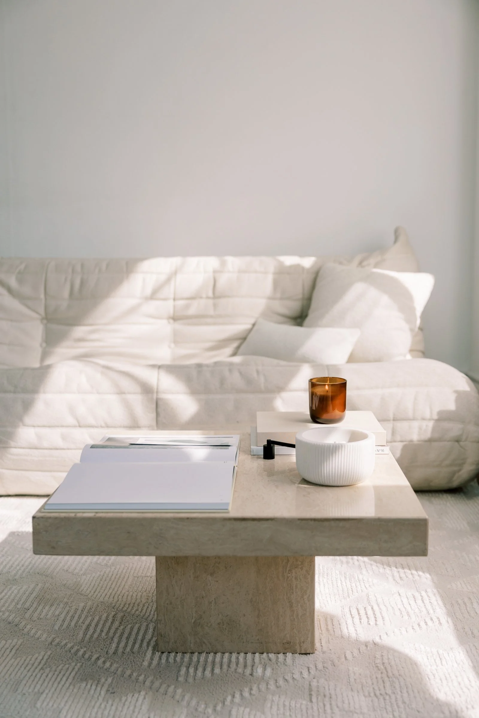 A beige marble coffee table with a white open book, a white ribbed bowl, a black pen, and a small black object on surface. Behind it, a white sofa with white pillows, a white blanket, and a amber glass candle holder. The background has a white wall.