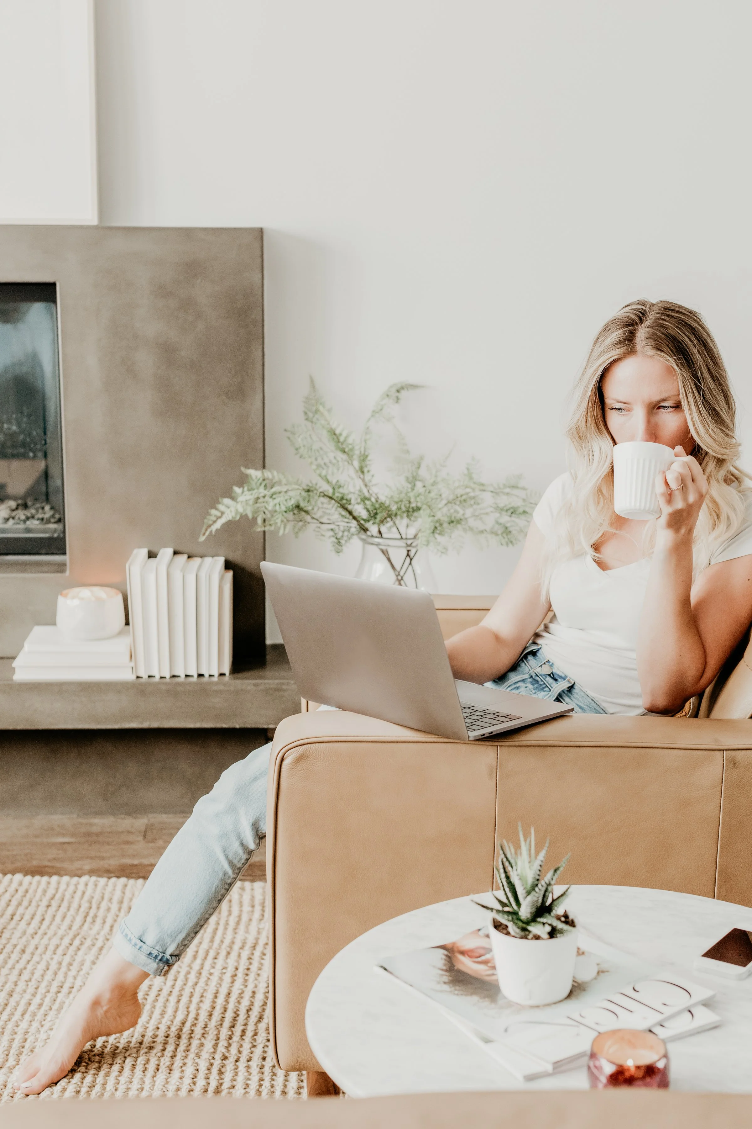 Young woman sitting on a beige armchair in a modern living room, sipping coffee from a white mug while working on a laptop. The room features a white side table with magazines, plants, and decor, and a beige carpet.
