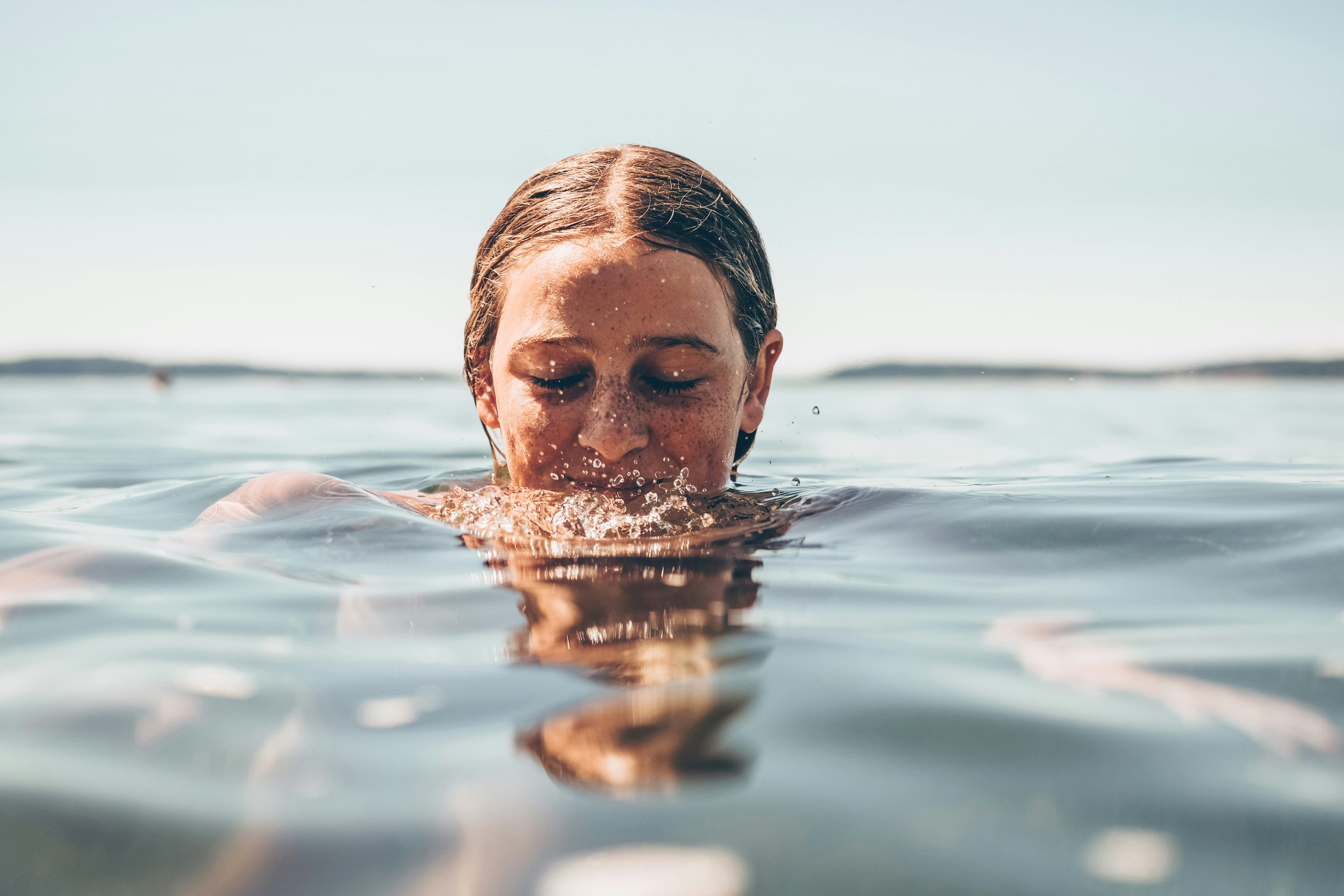 A girl with wet hair and closed eyes swimming in the water, with a clear sky and distant shoreline in the background.