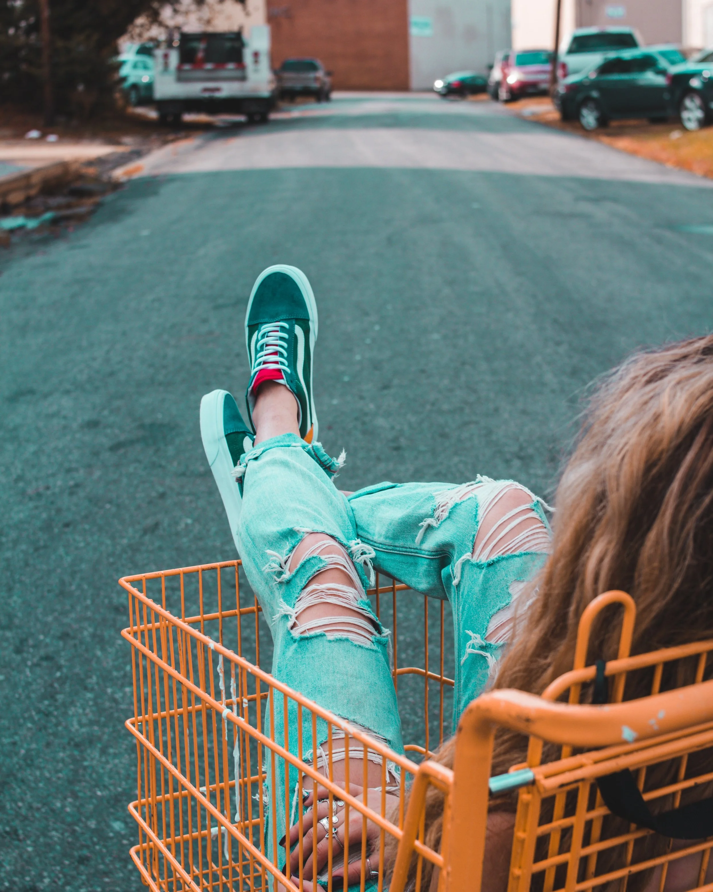 Young woman sitting in a shopping cart with legs extended, wearing ripped jeans and sneakers, on a street with parked cars in the background.