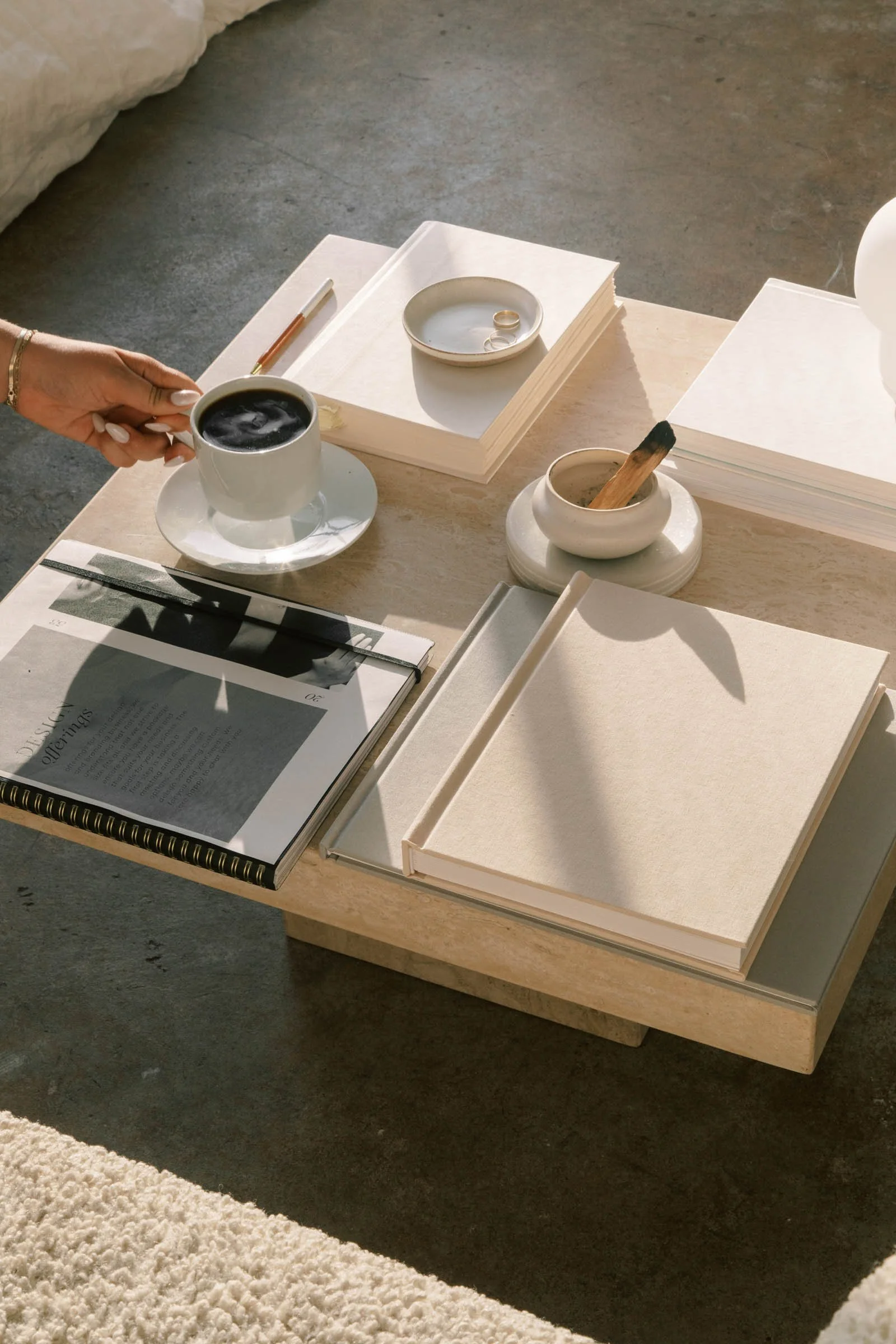 A wooden table holding a white ceramic mug of coffee, a stack of white papers, a notebook, a pen, a small bowl with a ring and a stick of incense, all in a softly lit room.