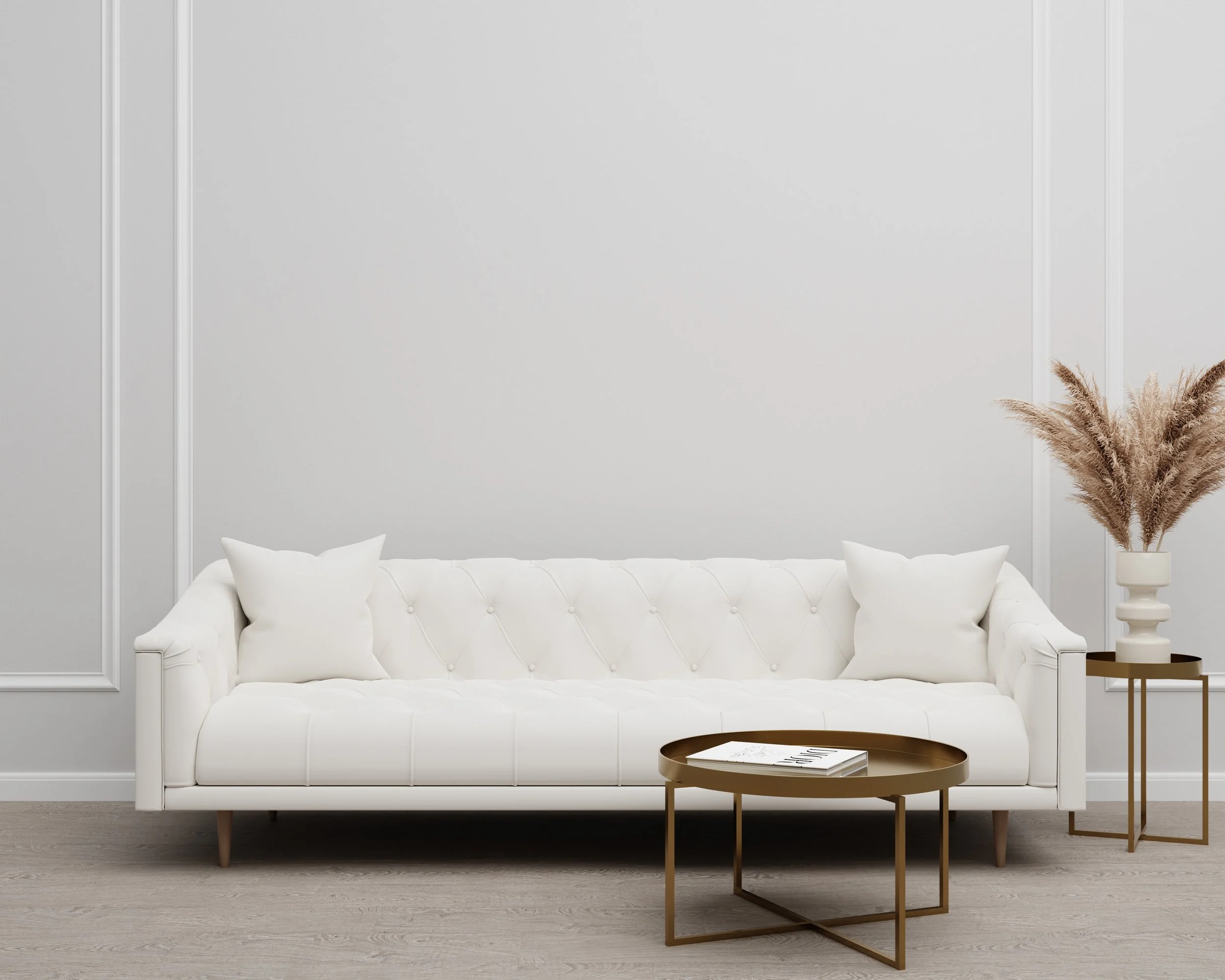 Minimalist living room with white tufted sofa, white walls, and simple decor. A round gold and brown coffee table with a magazine, a side table with a book, and a vase with beige pampas grass are visible.