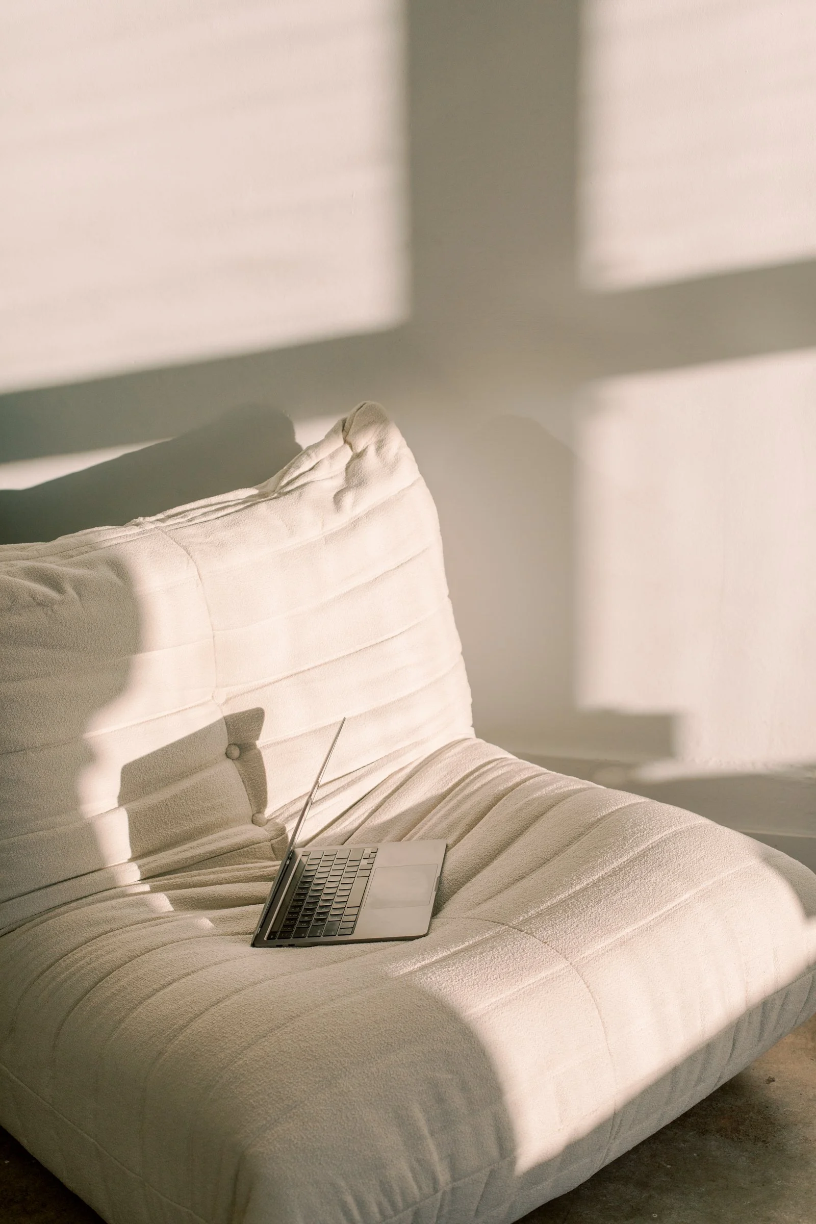 Close-up of a beige upholstered bed or couch with a laptop placed on it. Shadows of window frames are cast across the wall and furniture in a softly lit room.