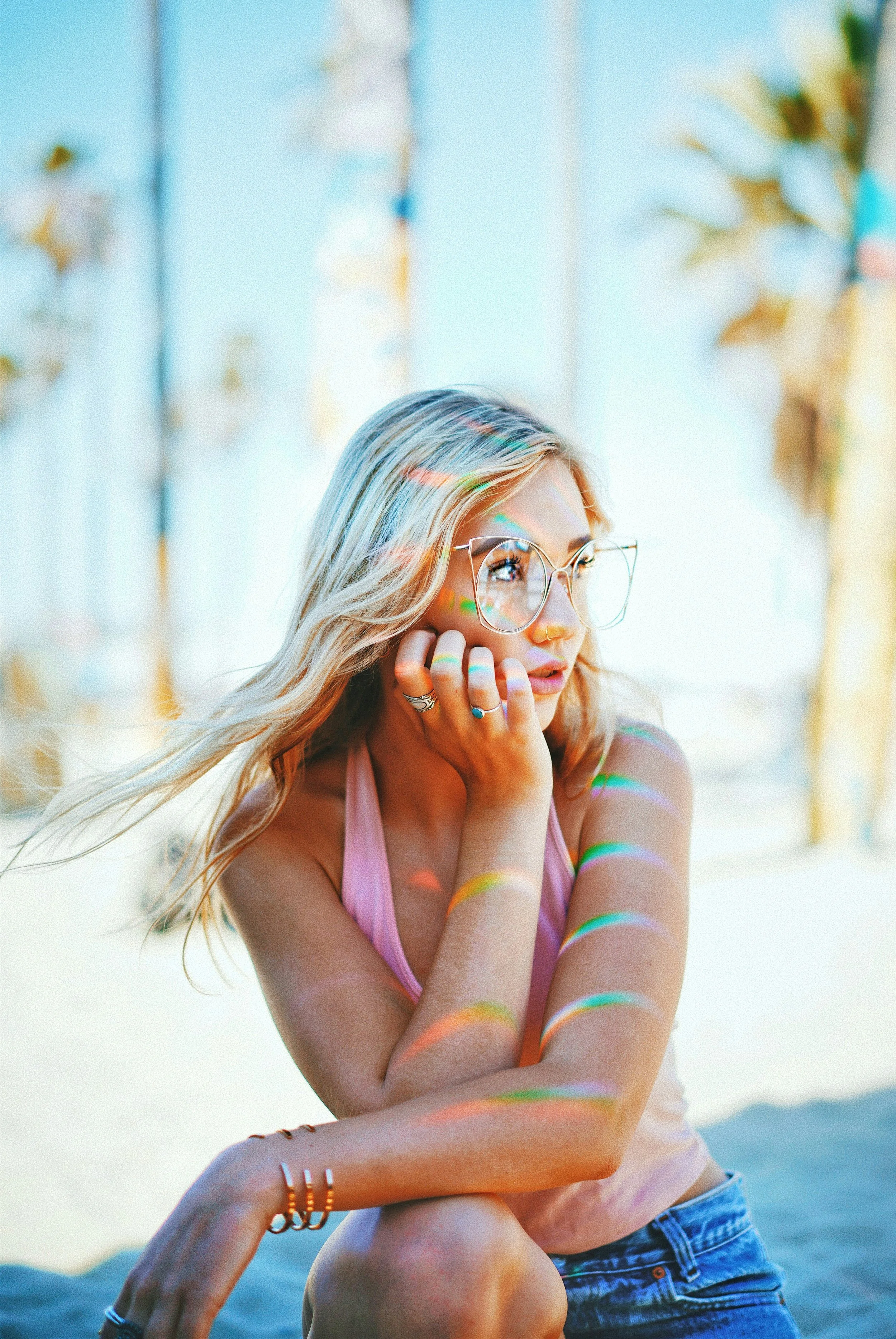 Young woman with sunglasses and blonde hair sitting outdoors with colorful light reflections on her skin.