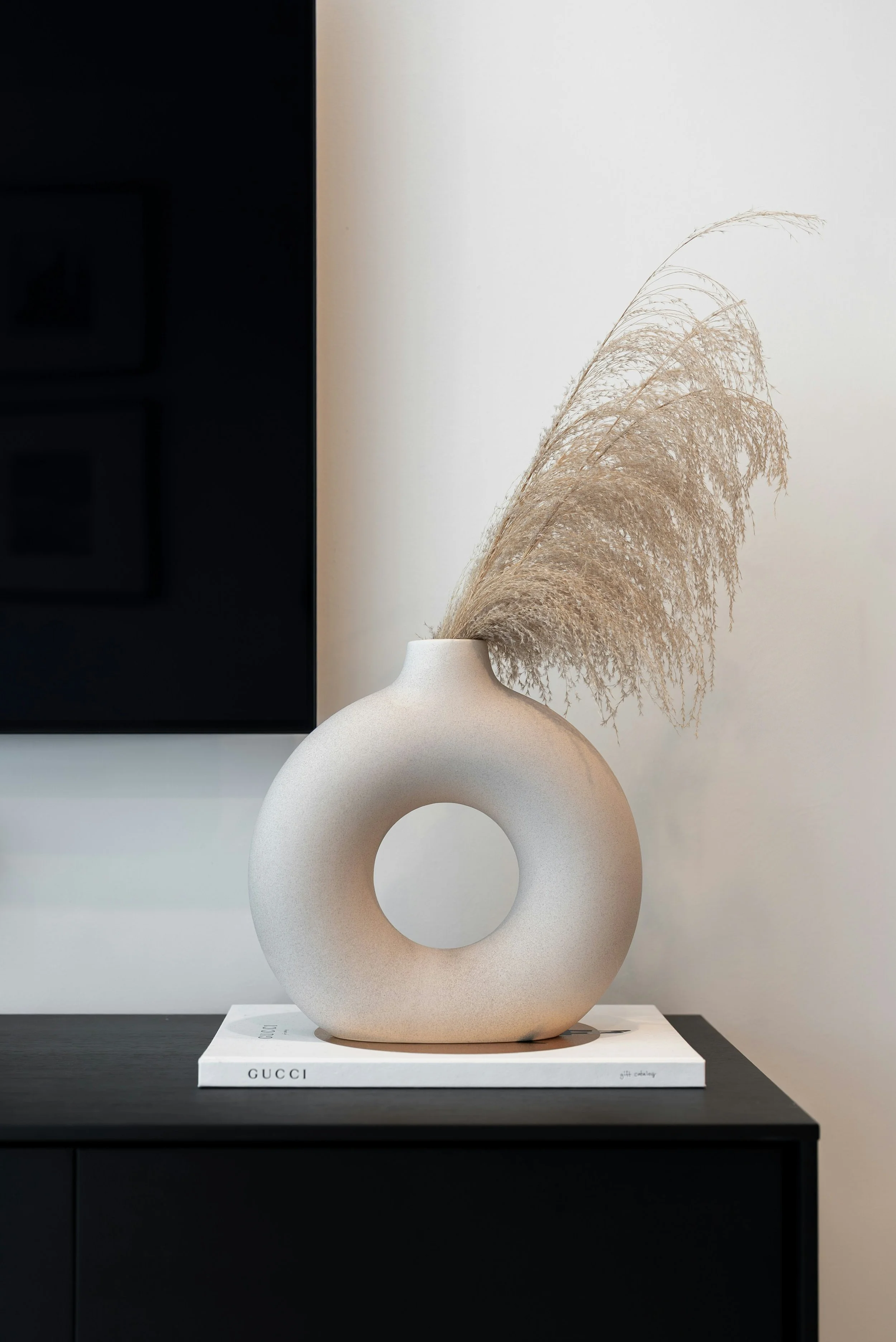 A beige ceramic vase with a circular hole in the center, holding dried pampas grass, placed on a white Gucci book on a black surface.