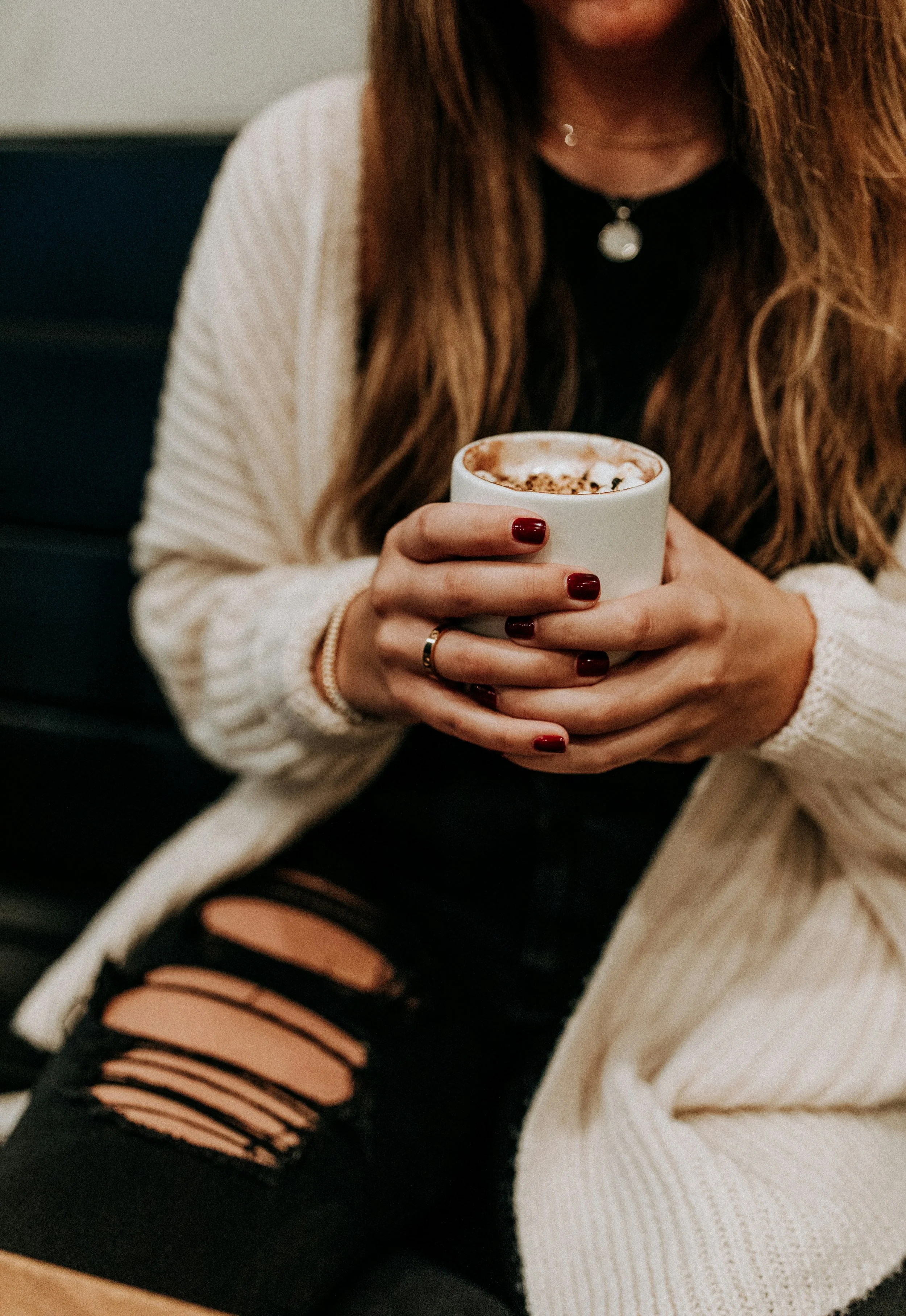 A female therapist with long hair wearing a cream-colored cardigan and black ripped jeans, holding a white mug with a hot beverage and whipped cream, sitting on a dark-colored couch.