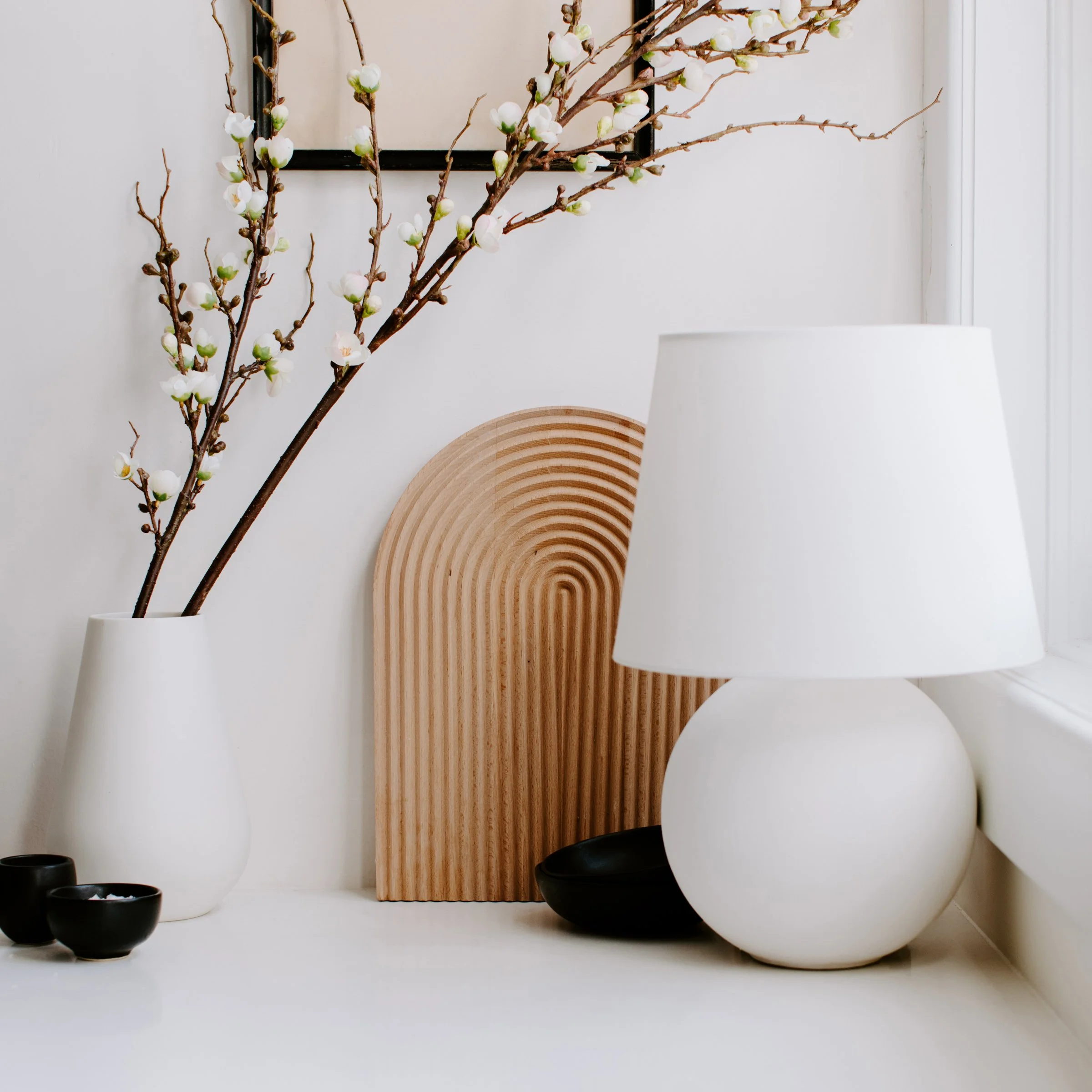Decorative indoor scene with vases, a piece of curved wood art, and bowls, arranged on a white surface against a white wall.