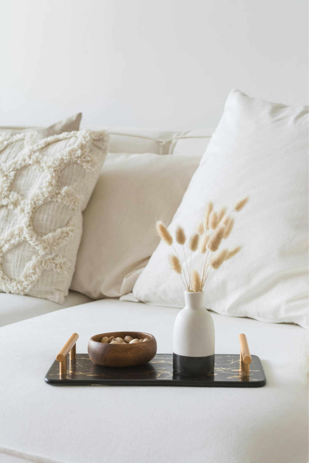 A cozy, neutral-toned living room with a beige sofa, textured throw pillow, and a black marble tray on the sofa's surface. The tray holds a wooden bowl with nuts and a modern white vase with dried beige bunny tail grass.