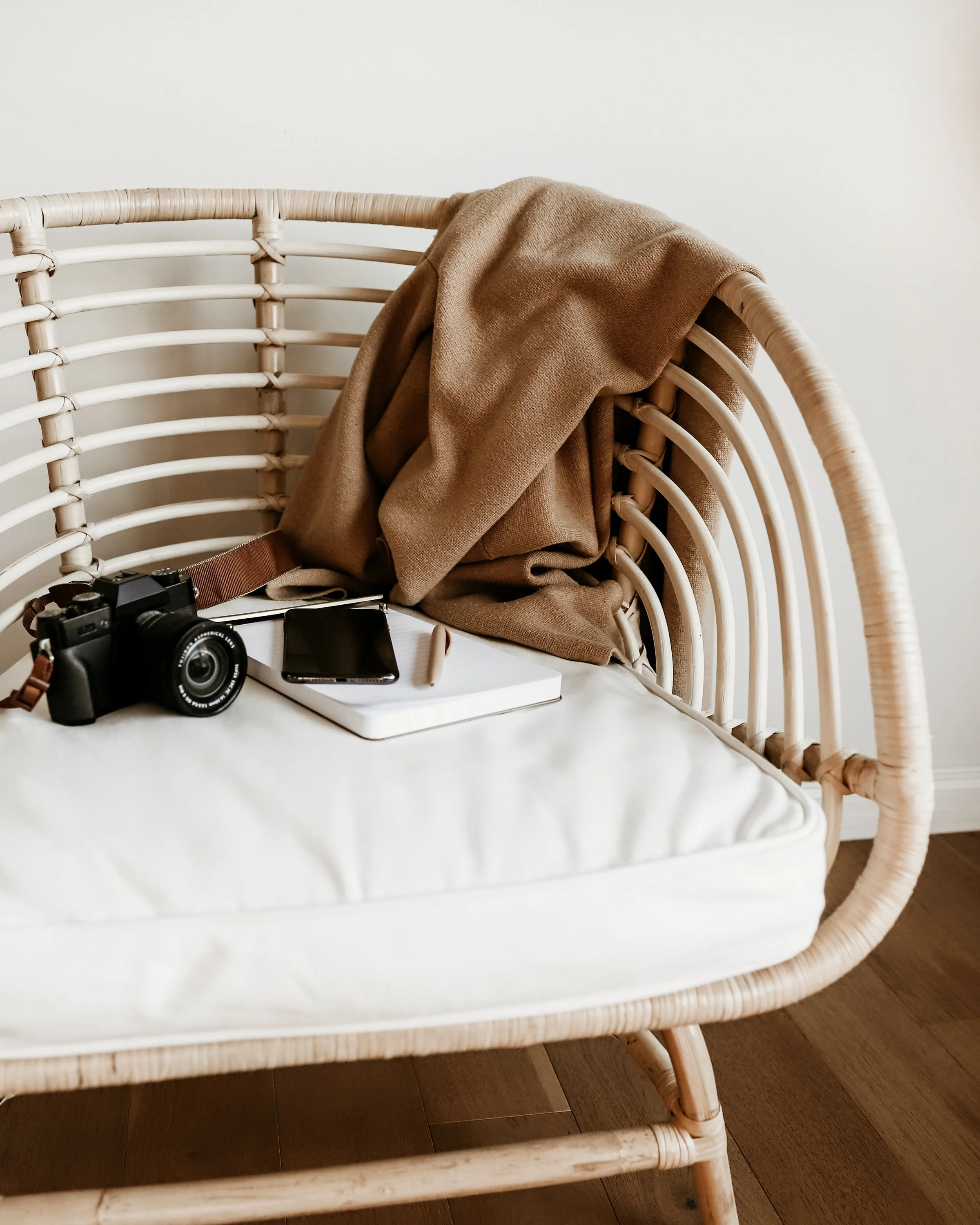 A rattan chair with a beige blanket draped over the backrest, a camera, a smartphone, a book, and a pen placed on a white surface, against a plain wall.