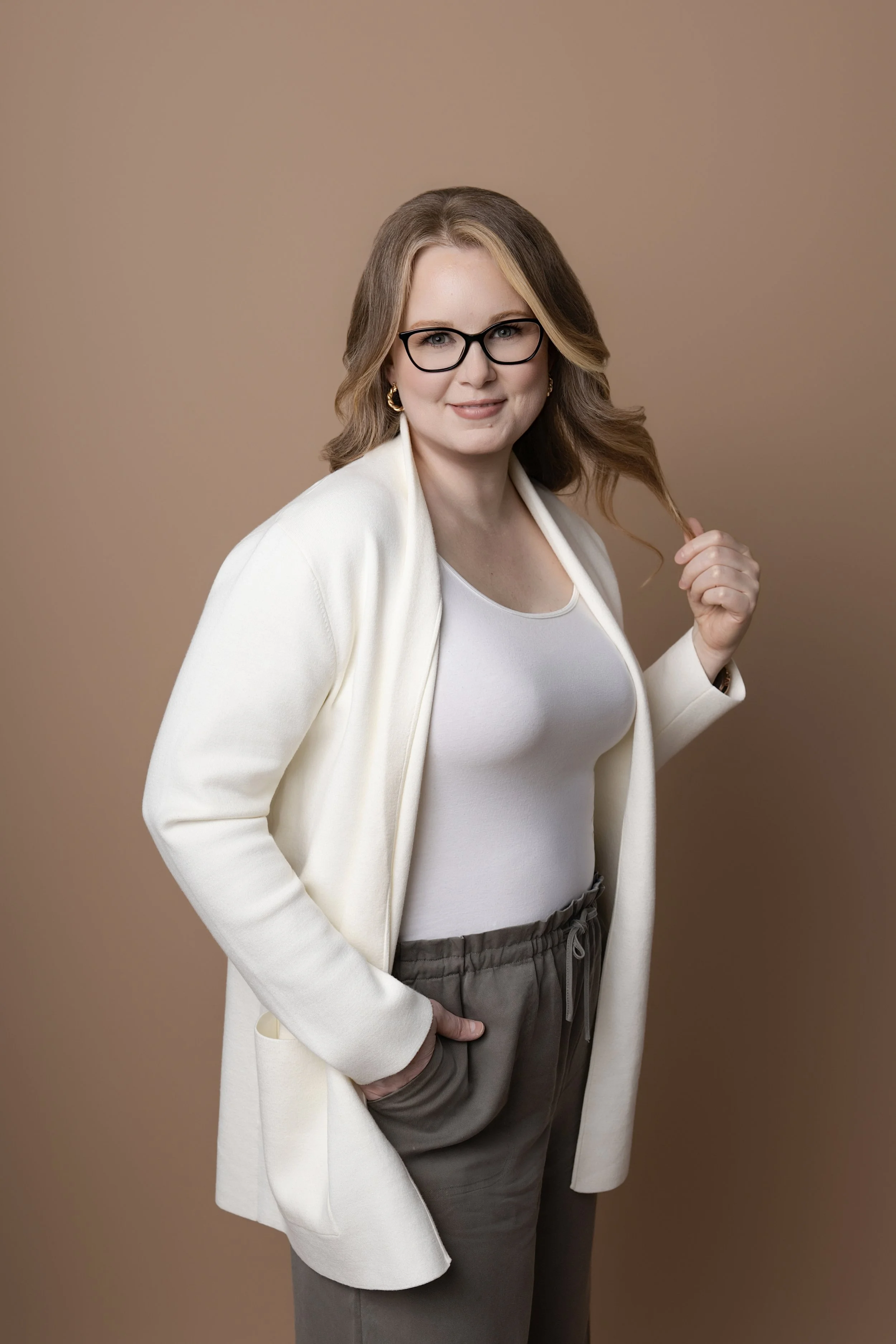 Therapist Julie Thomason with wavy light brown hair, wearing glasses, a white blazer over a white top, and gray pants, is standing against a beige background. She is smiling and holding a lock of her hair with her right hand.