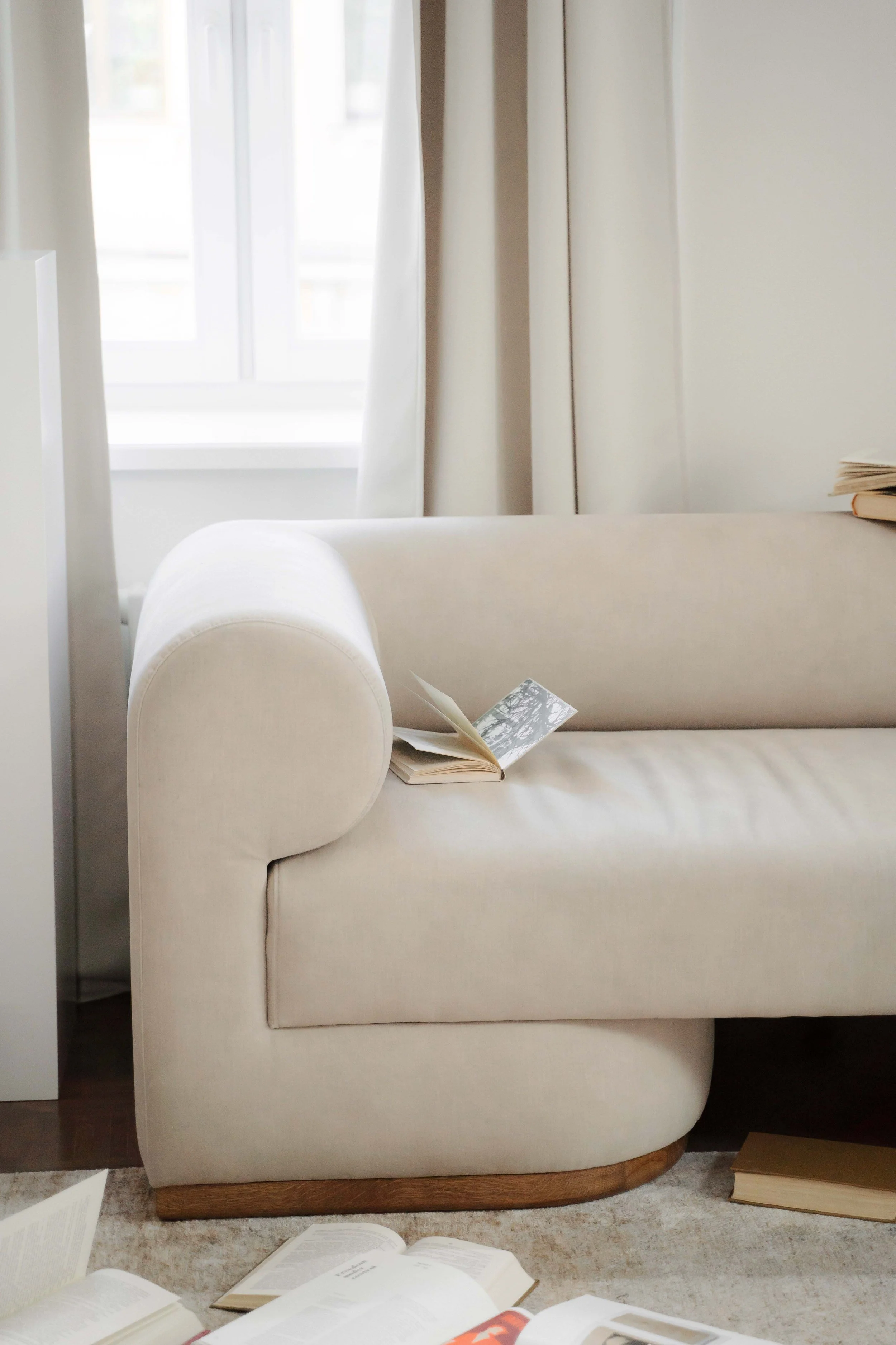 A cream-colored upholstered armchair with a rounded armrest and wooden base, placed on a beige carpet. An open book rests on the seat, and several other books and papers are scattered on the floor and the armrest. Light filters through a window with white curtains in the background.