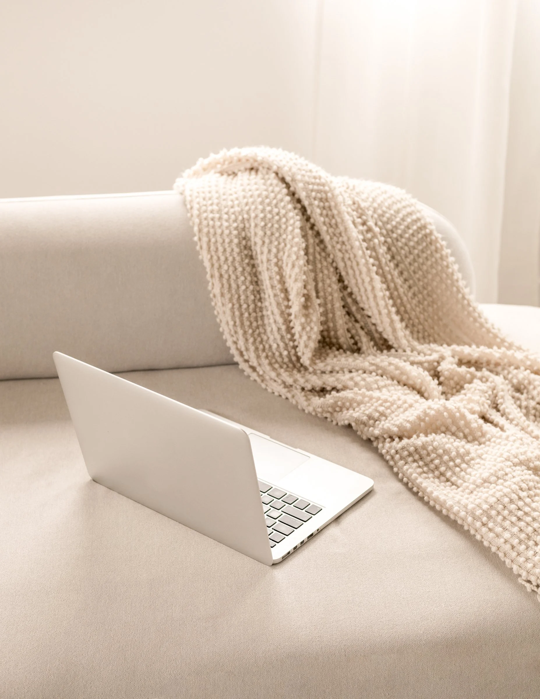 A beige knitted blanket draped over the back of a light gray sofa, with a silver laptop sitting on a white table in the foreground.
