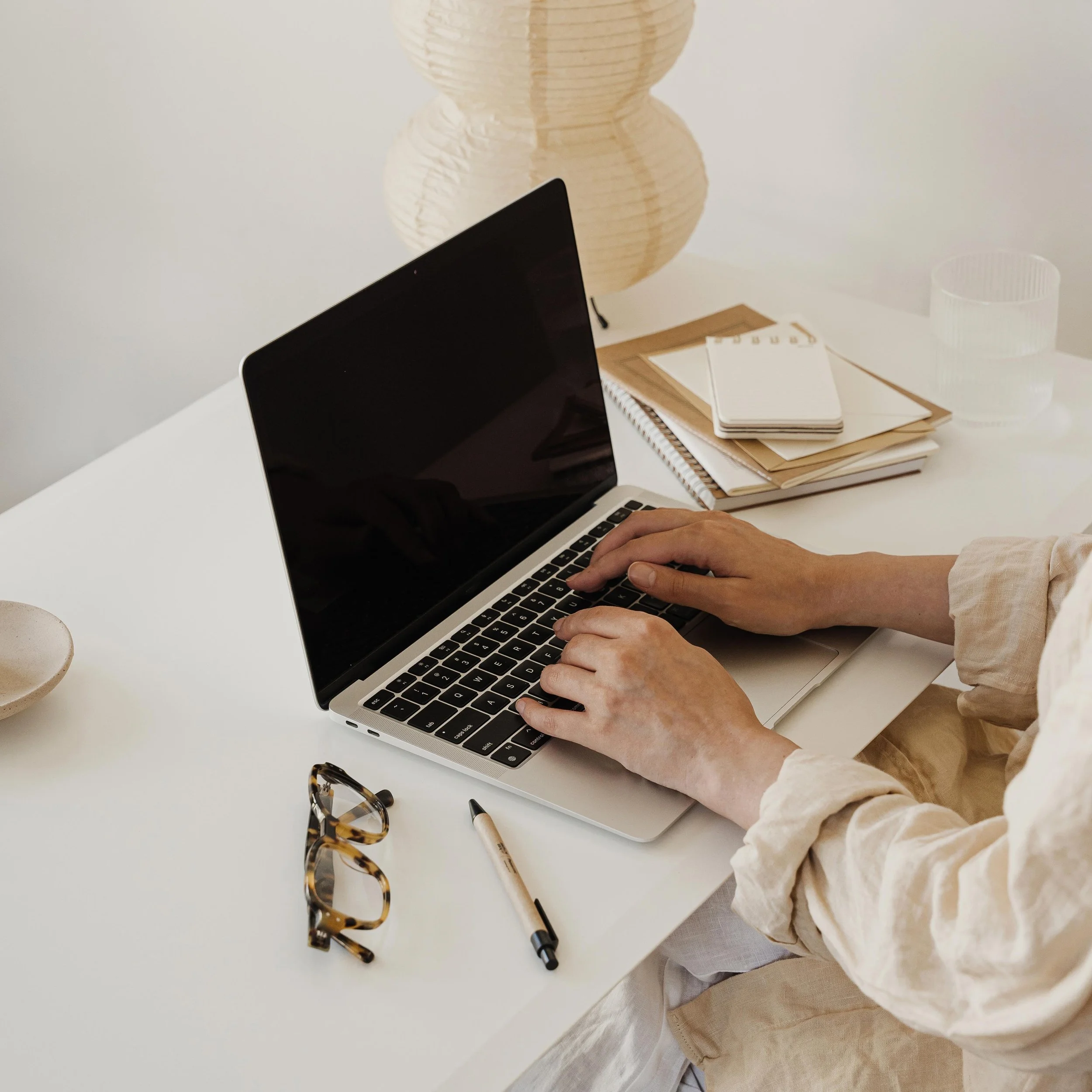 A therapist at a desk, typing on a laptop with glasses and a notebook.