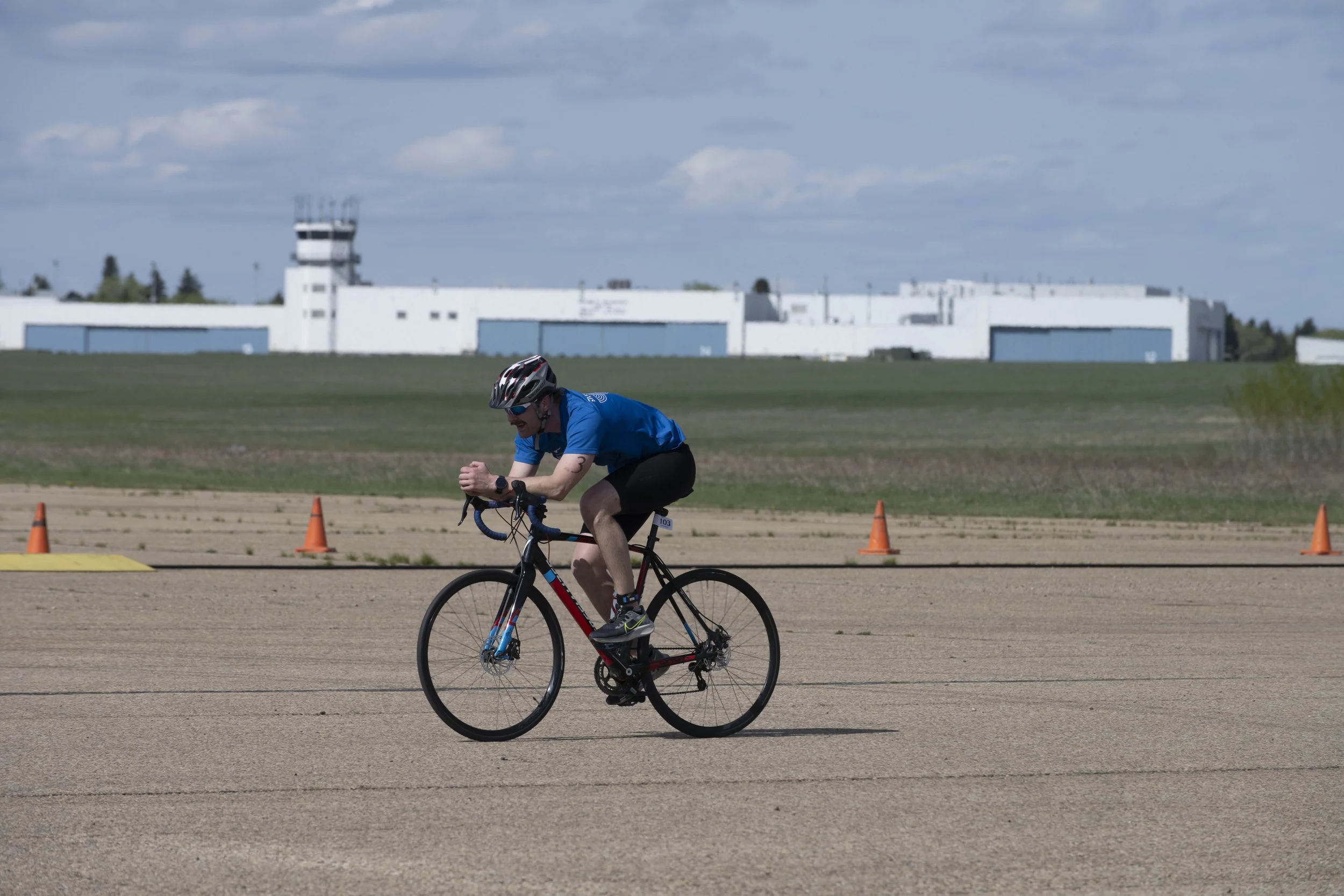 A person wearing a blue shirt, black shorts, a helmet, and sunglasses riding a bicycle on a flat paved surface, with orange traffic cones and an airport terminal with a control tower in the background under a partly cloudy sky.