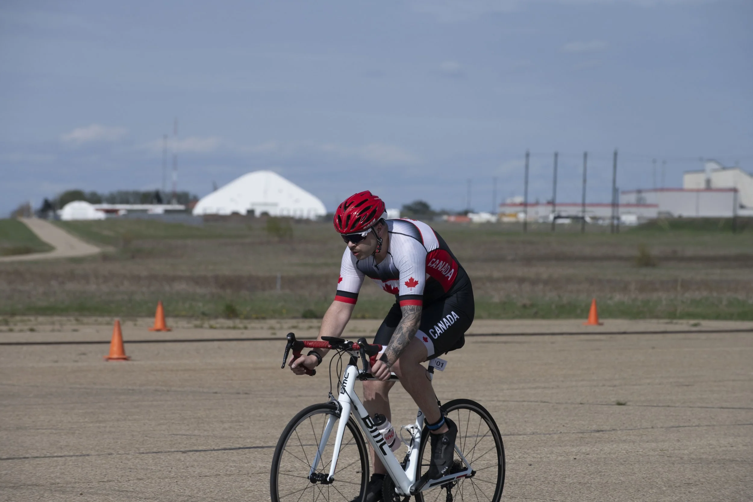 A male cyclist wearing a red helmet, sunglasses, and a Canada-themed cycling outfit riding a white BMC bicycle on a flat, open track with orange cones and industrial buildings in the background.